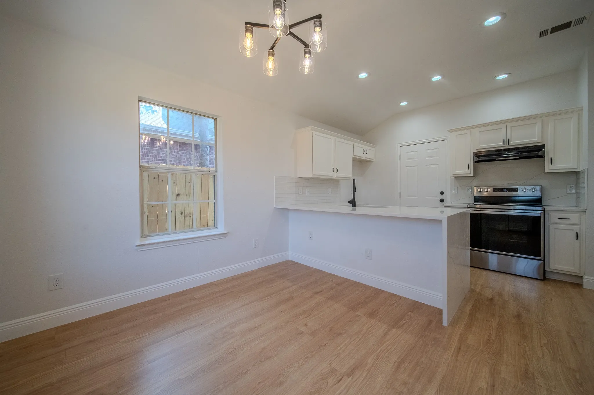 Open-concept living space featuring wood-finish flooring, a modern linear chandelier, recessed lighting, and a kitchen with white cabinetry, a subway tile backsplash, and stainless steel appliances