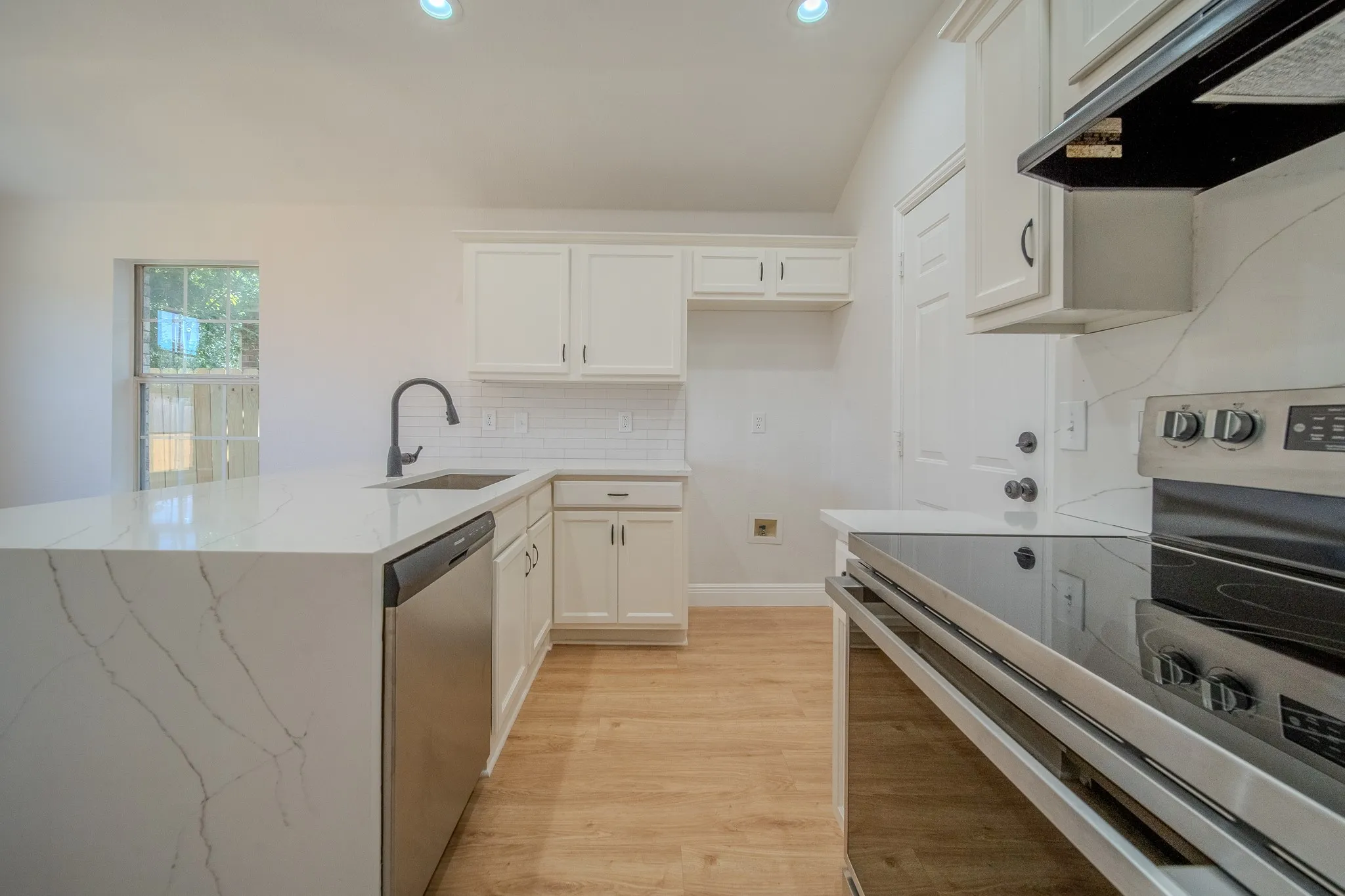 Contemporary kitchen featuring a waterfall-edge island with quartz countertops, stainless steel appliances, a matte black gooseneck faucet, white shaker cabinetry, and wood-finish flooring