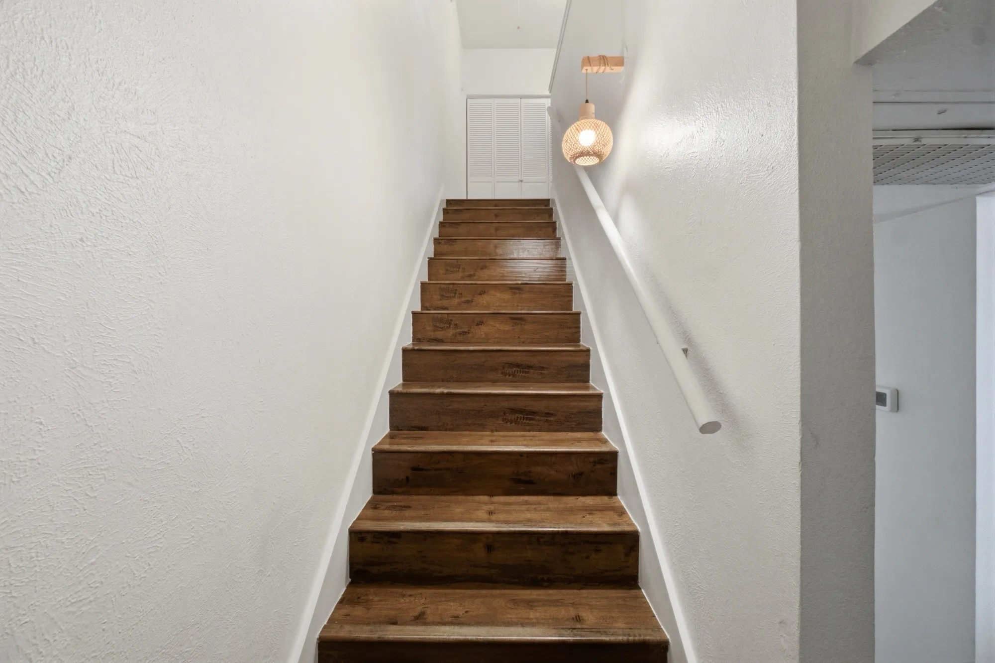 Wood-finish staircase with a white handrail, smooth white walls, and a decorative pendant light fixture