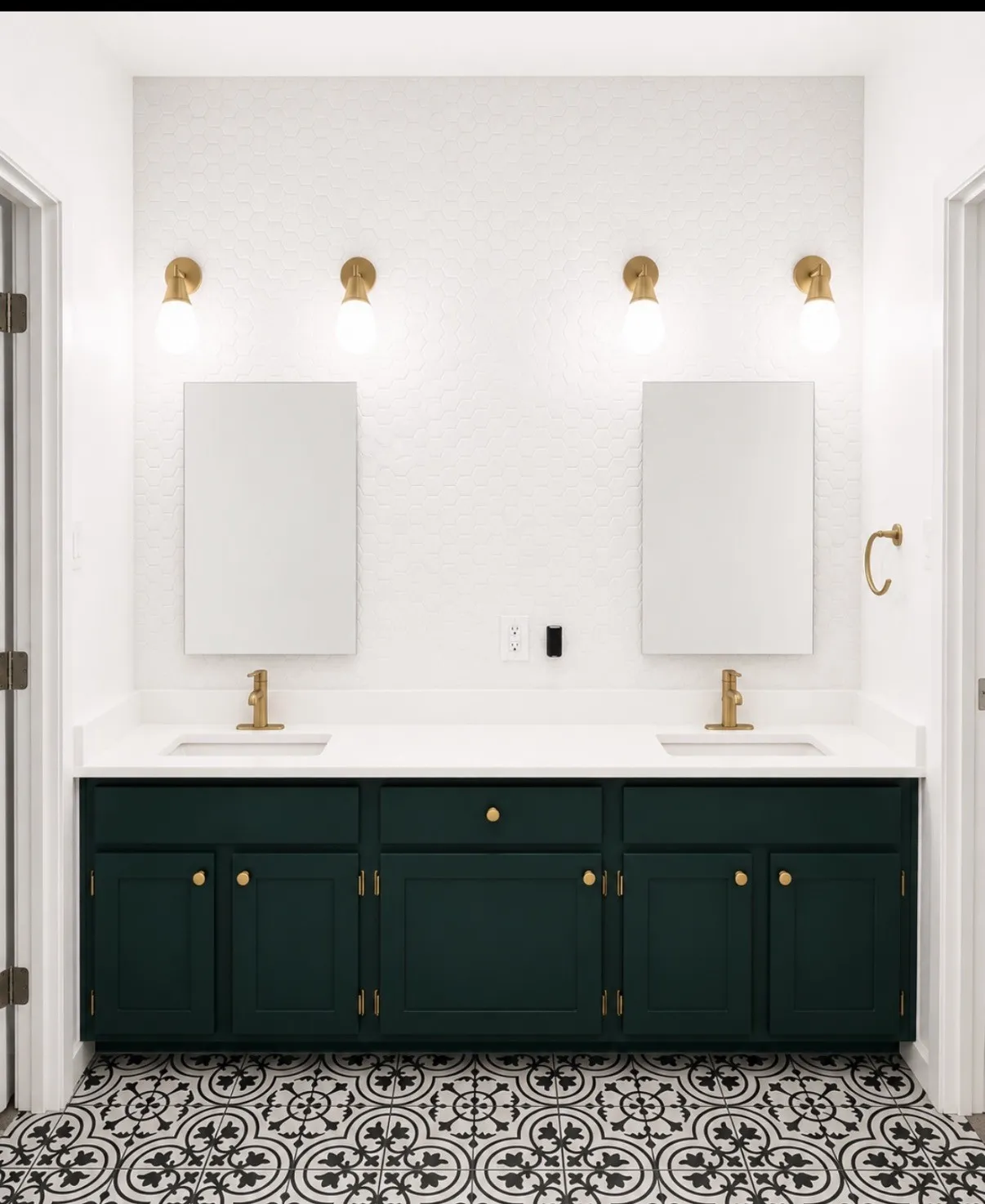 Double vanity with a white countertop and dark green cabinetry, featuring dual rectangular sinks with gold-finish faucets, overhead wall-mounted lighting, and patterned floor tile