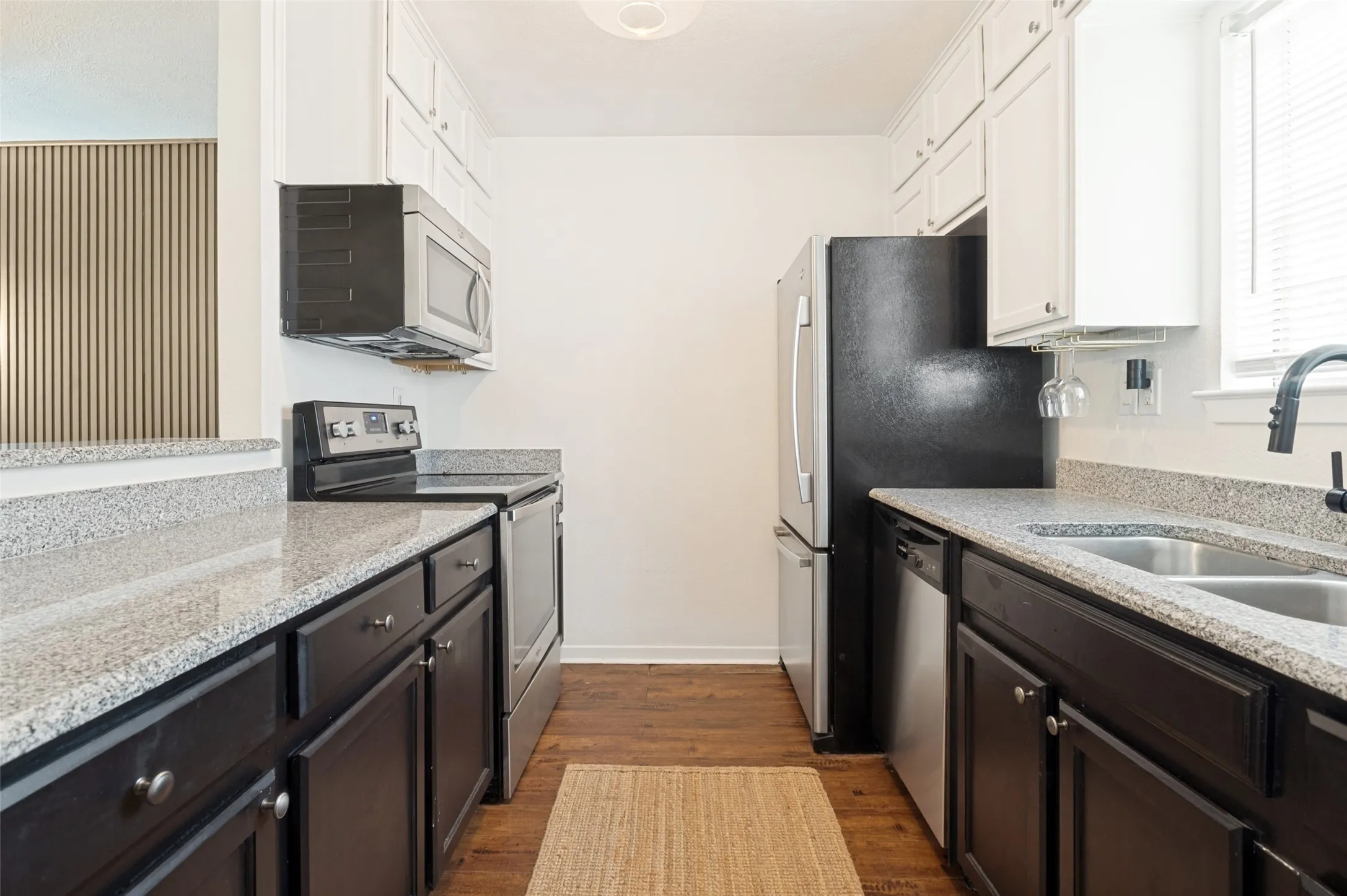 Galley kitchen featuring granite countertops, stainless steel appliances, wood-finish flooring, white upper cabinetry, and dark lower cabinetry