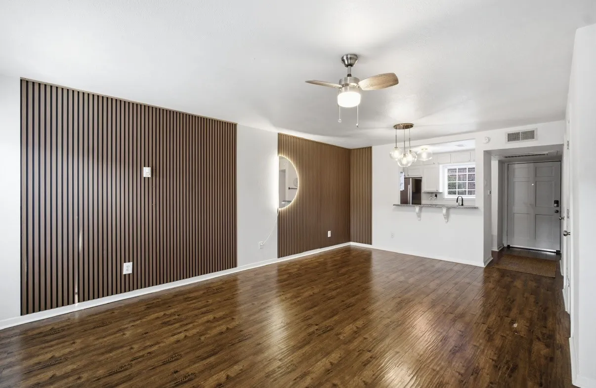Wood-finish flooring, accent wall with vertical slats, ceiling fan with integrated lighting, kitchen pass-through with counter, and pendant lighting