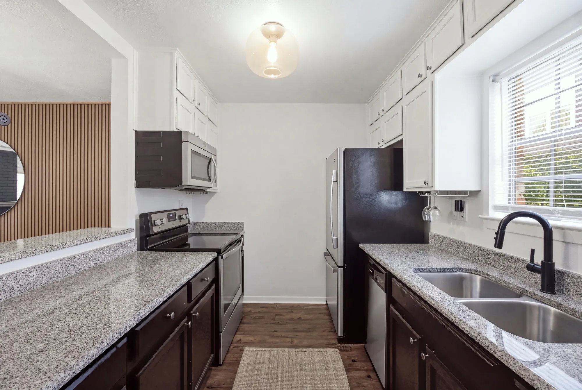 Galley kitchen featuring granite countertops, wood-finish flooring, white upper cabinetry, dark lower cabinetry, and a double basin stainless steel sink with a matte black faucet