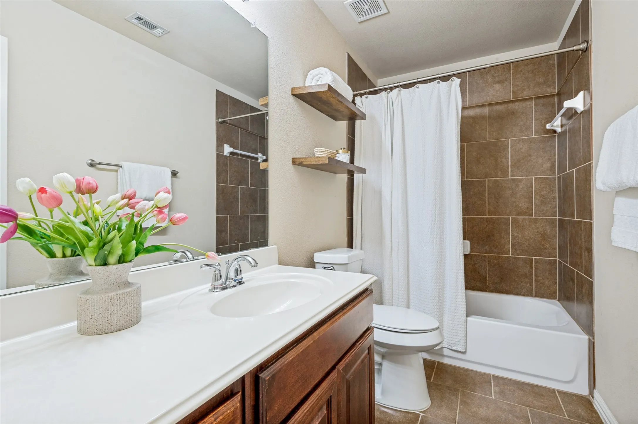 Bathroom featuring a brown tiled bath and shower combination, a white countertop vanity with a chrome faucet, and wood-finish floating shelves