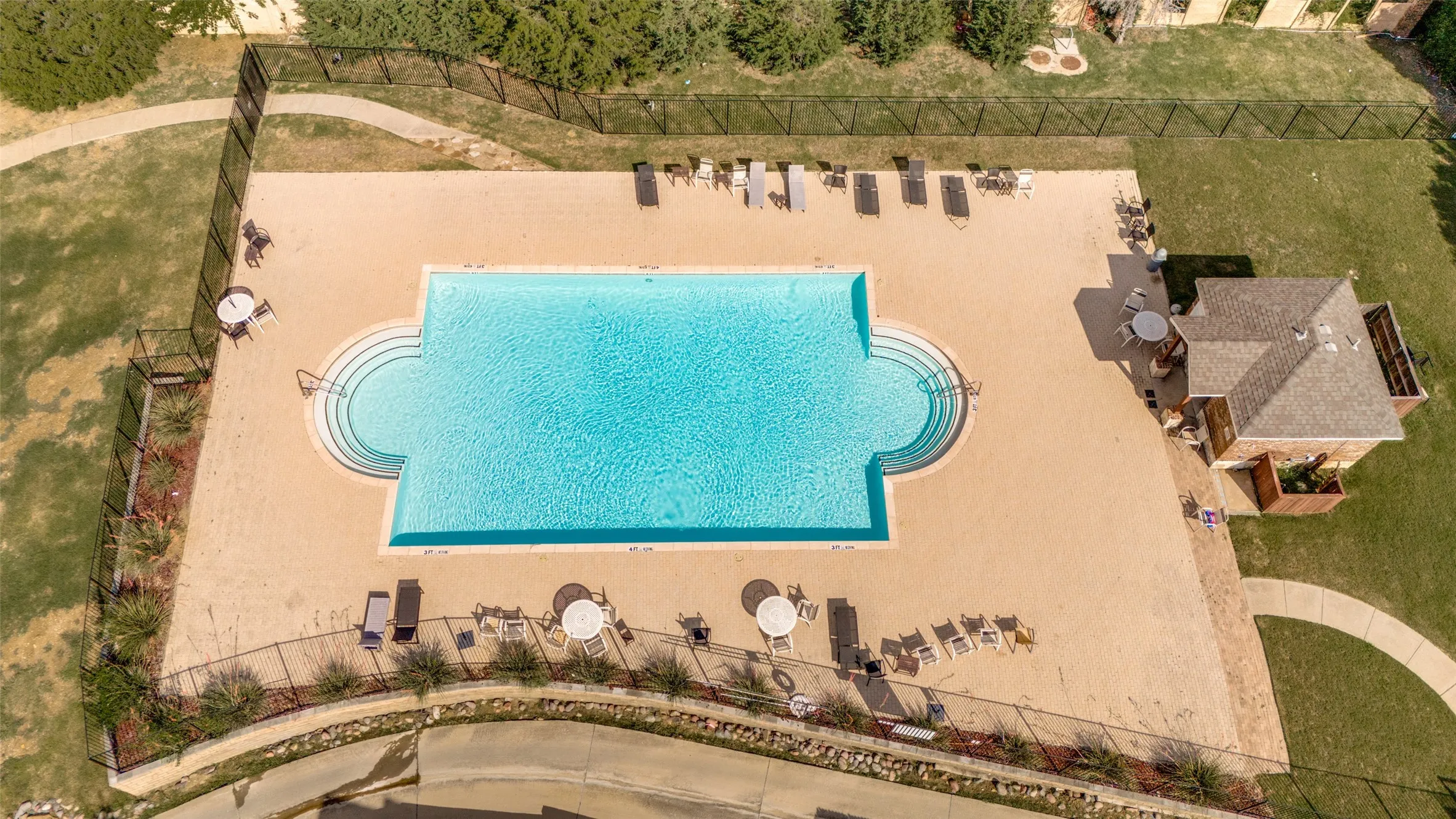 Community swimming pool featuring a perimeter fence, surrounding paved deck, and adjacent stone-clad cabana with a shingled roof