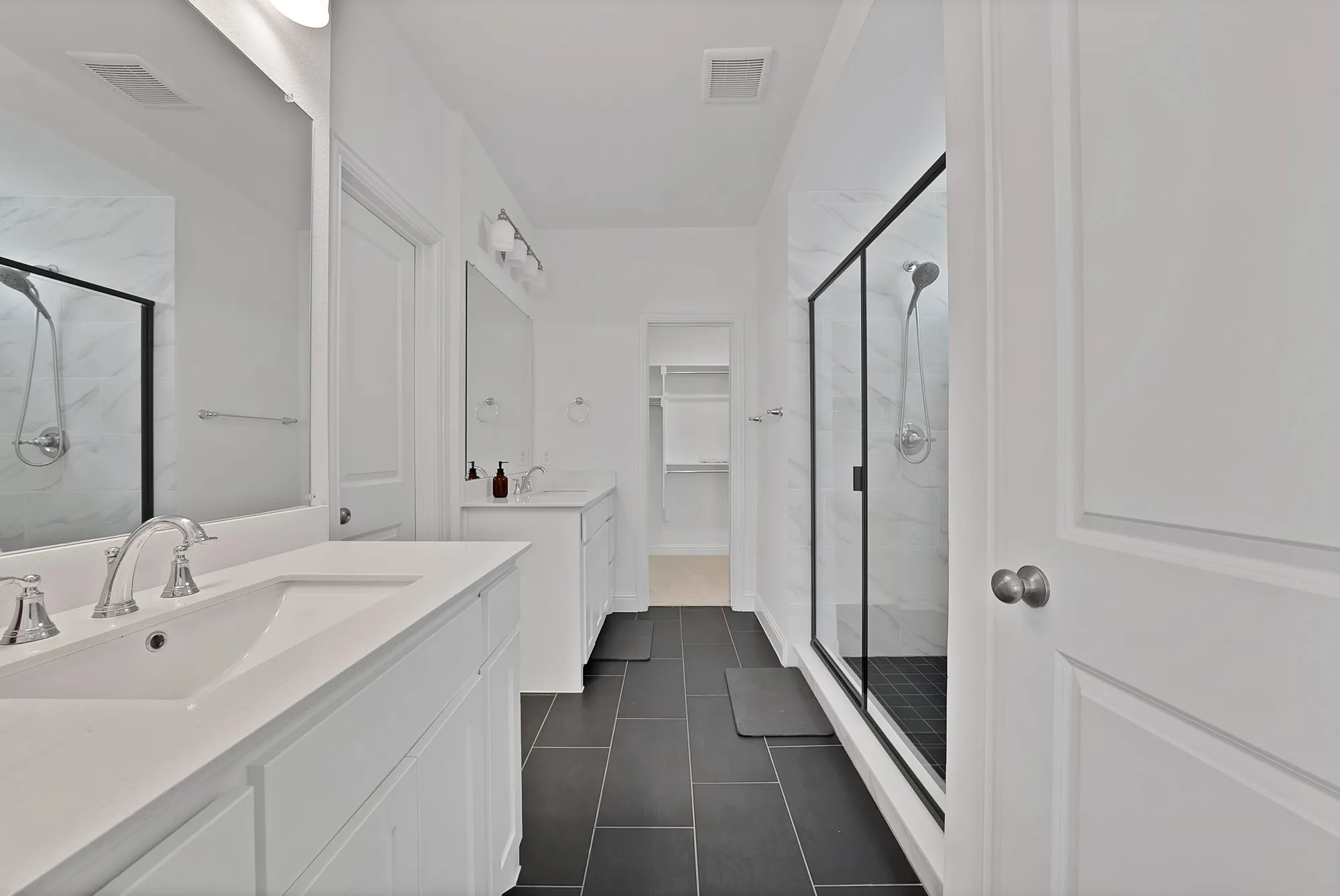 Spacious bathroom featuring dual white vanities with integrated sinks, brushed nickel fixtures, a large frameless mirror, and modern tile flooring