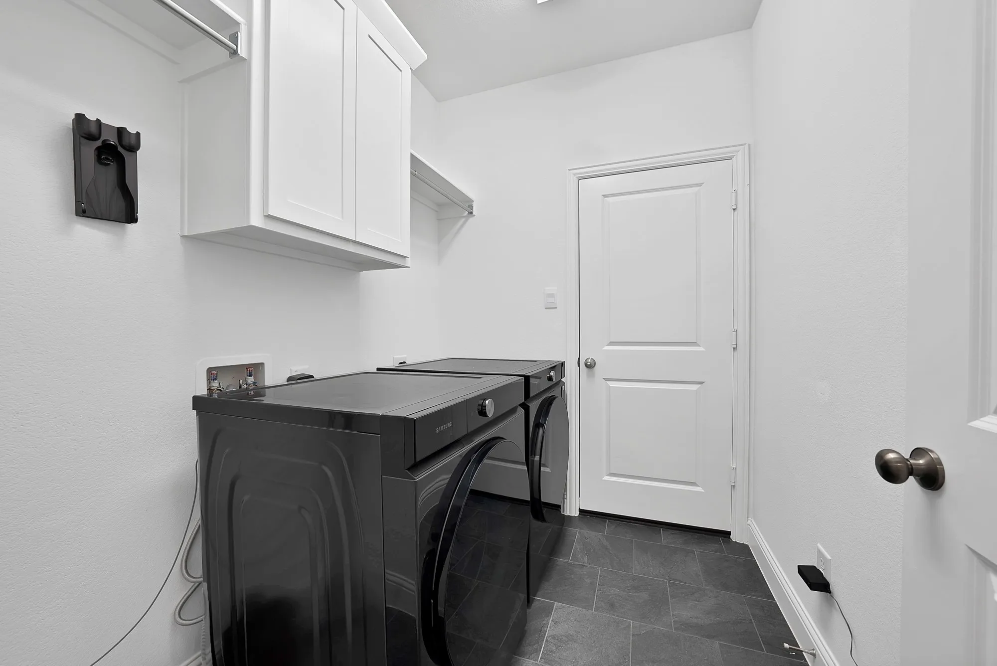 Laundry room featuring dark gray tile flooring, crisp white walls, white shaker-style cabinetry, and a wall-mounted shelving unit