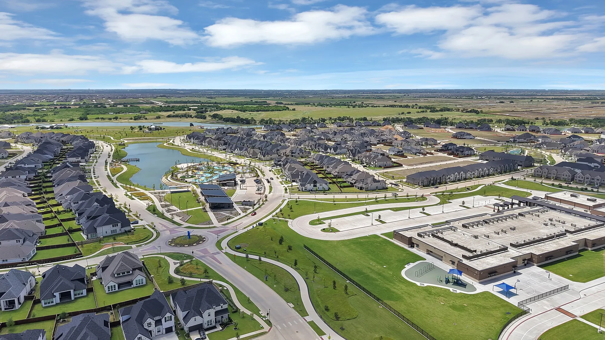 Expansive residential community featuring a central lake, extensive green spaces, a large school building, multiple recreational amenities, and curvilinear roadways