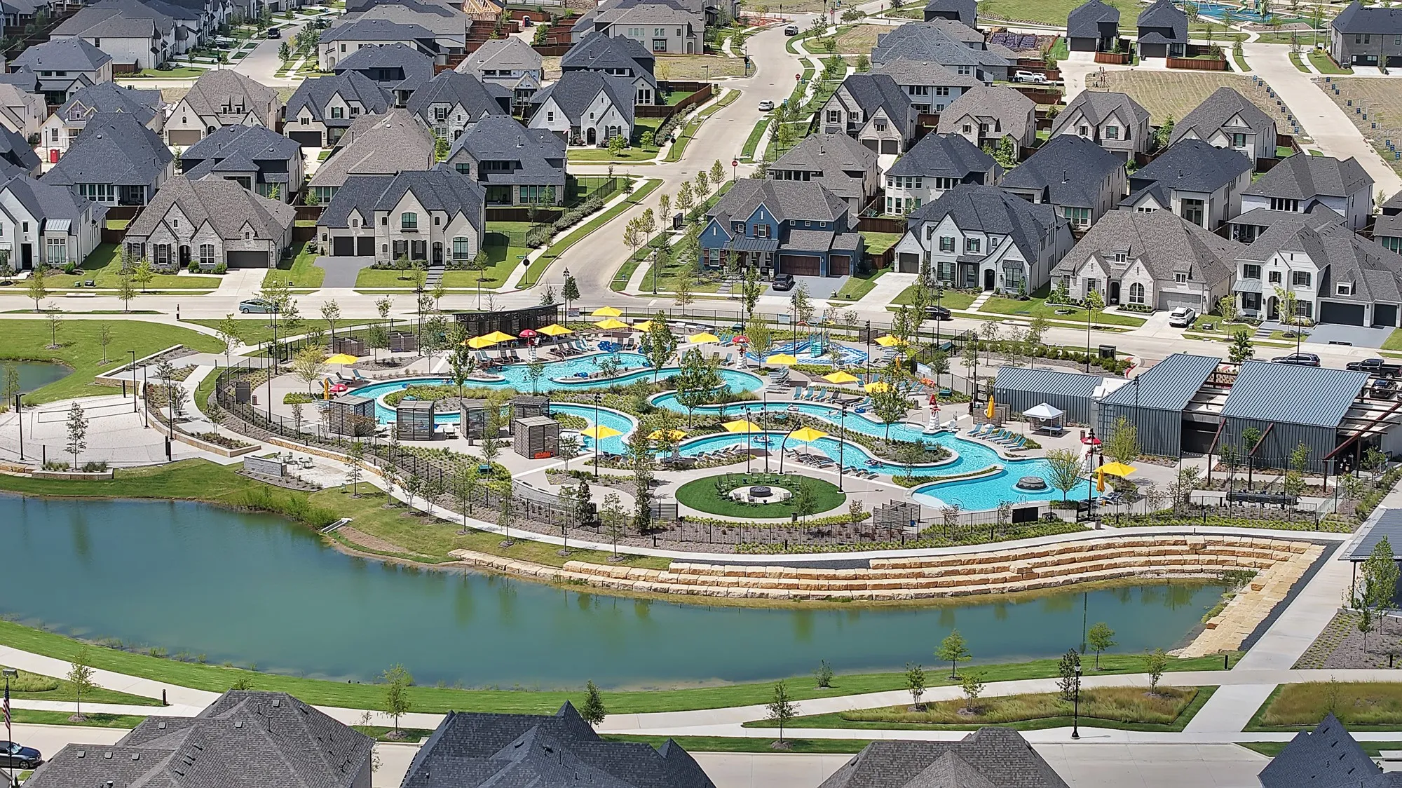 Expansive community featuring numerous residential properties with diverse architectural styles, a central water park with multiple pools and slides, a tranquil pond, and extensive manicured green spaces