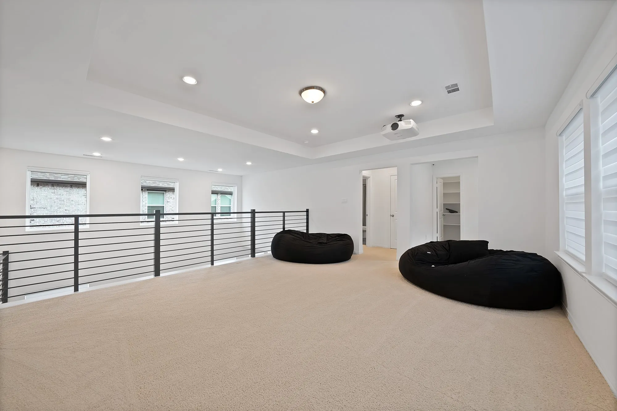 Carpeted loft area featuring a tray ceiling with recessed lighting, built-in projector, white window blinds, and contemporary horizontal bar railing