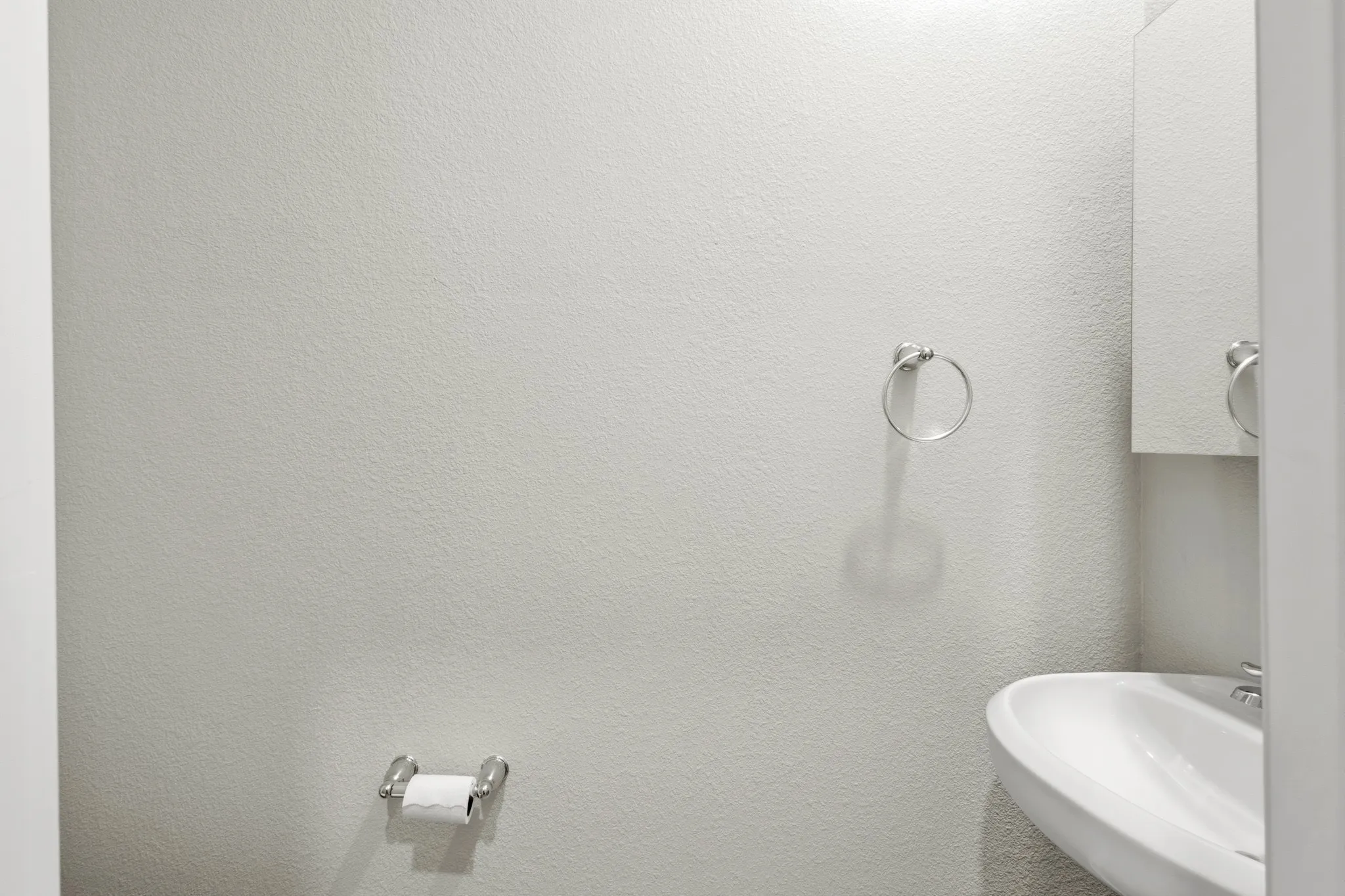 Wall-mounted ceramic sink with chrome faucet, complemented by a mirrored medicine cabinet, chrome towel ring, and chrome toilet paper holder