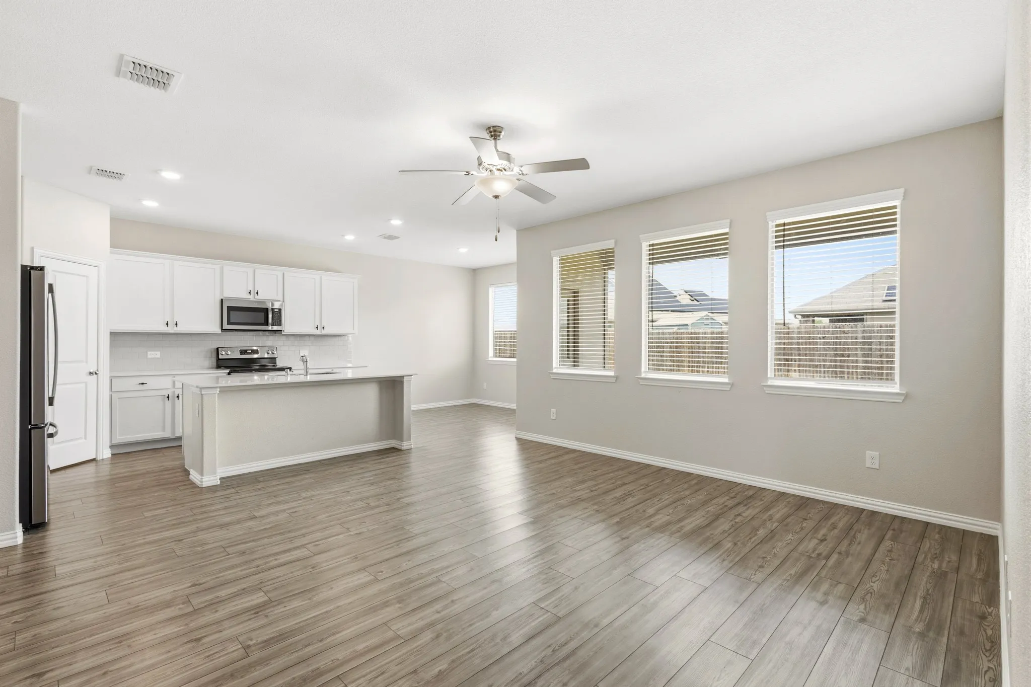 Open-concept space featuring wood-finish flooring, a kitchen island with a built-in sink, white cabinetry with stainless steel appliances, and multiple windows with blinds