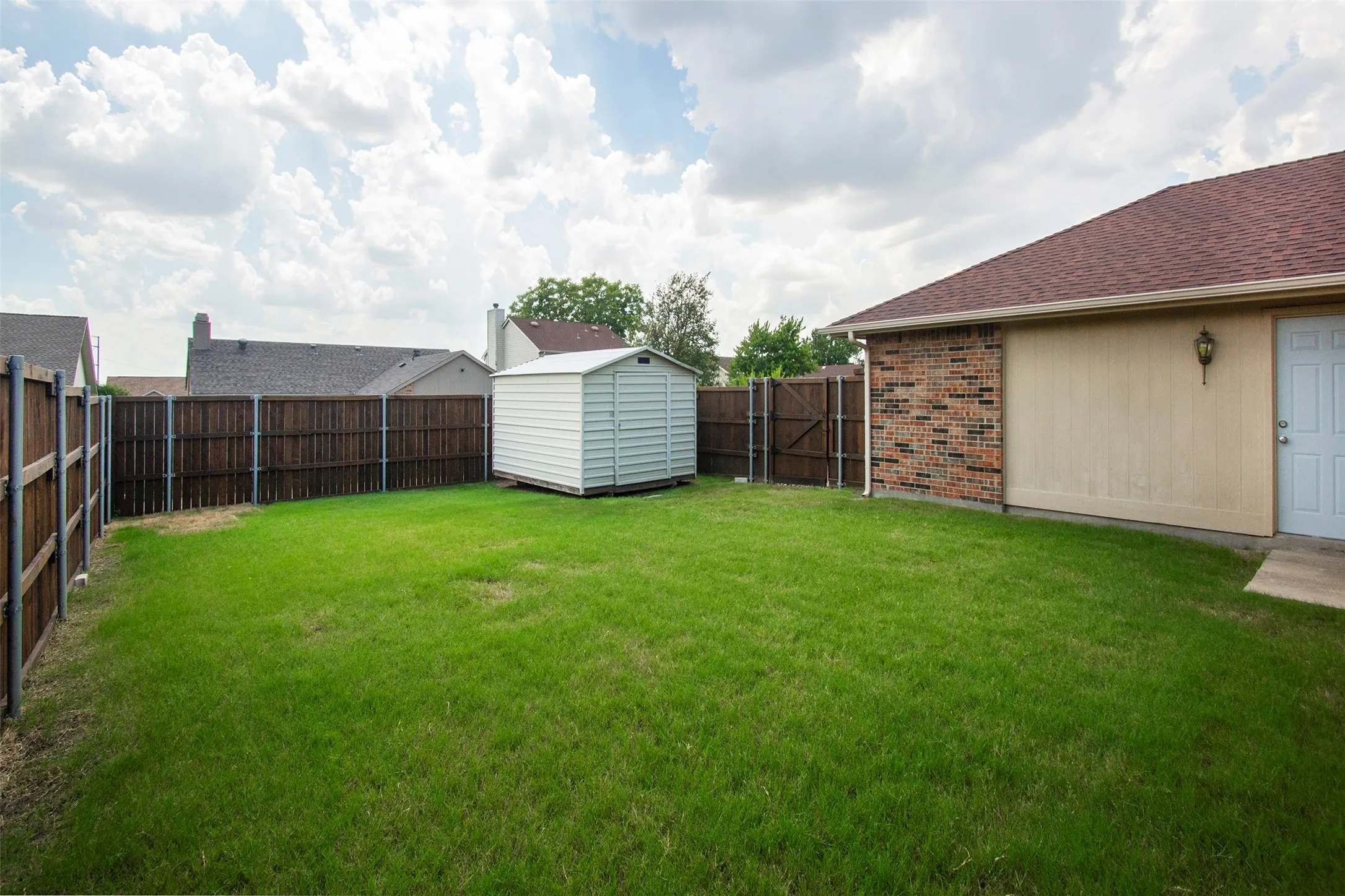 Expansive grassy yard featuring a wood privacy fence, a detached storage shed, and a brick and siding exterior wall with an exterior door