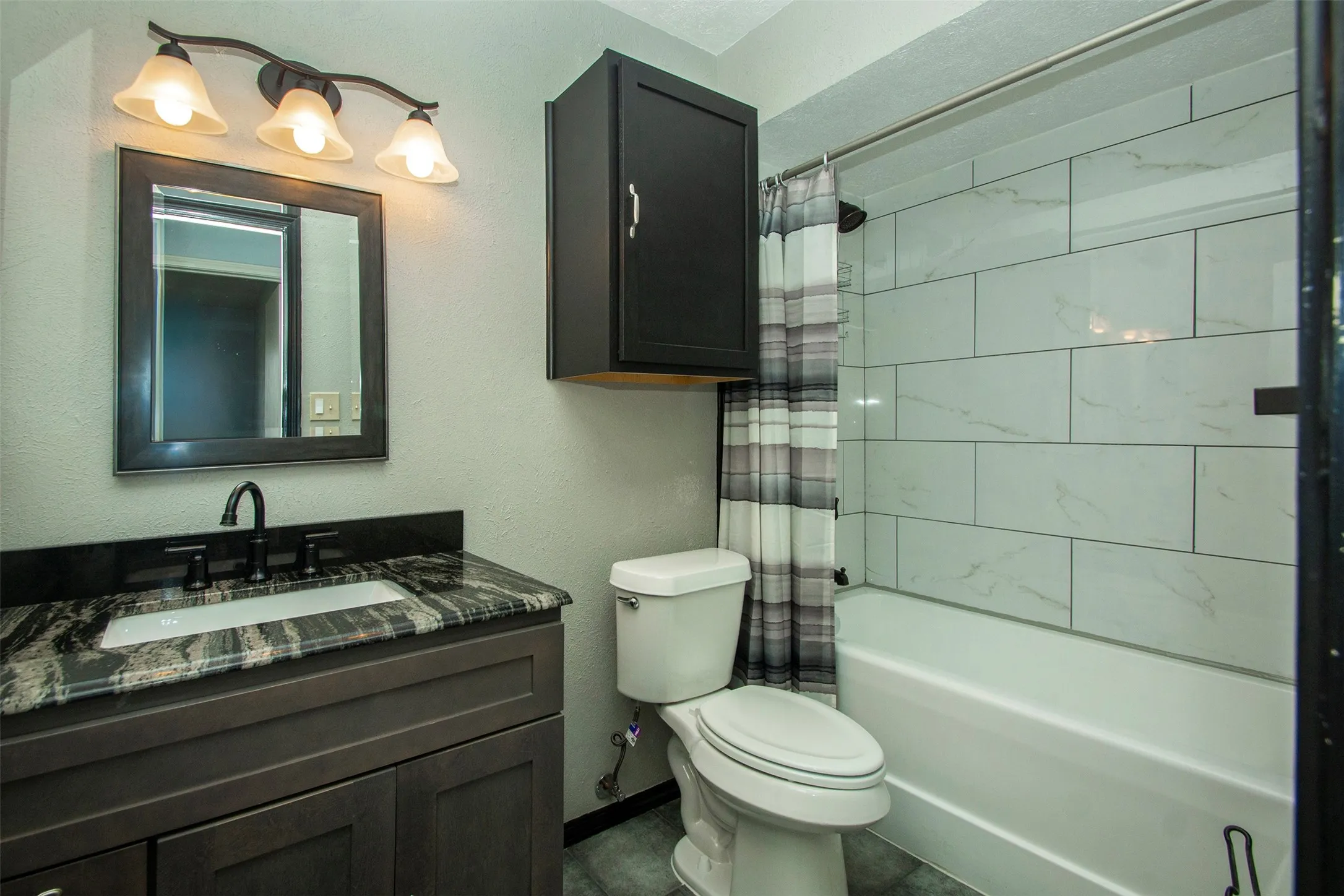 Bathroom featuring a dark wood-finish vanity with an undermount sink, a patterned countertop, black fixtures, and subway tile shower surround