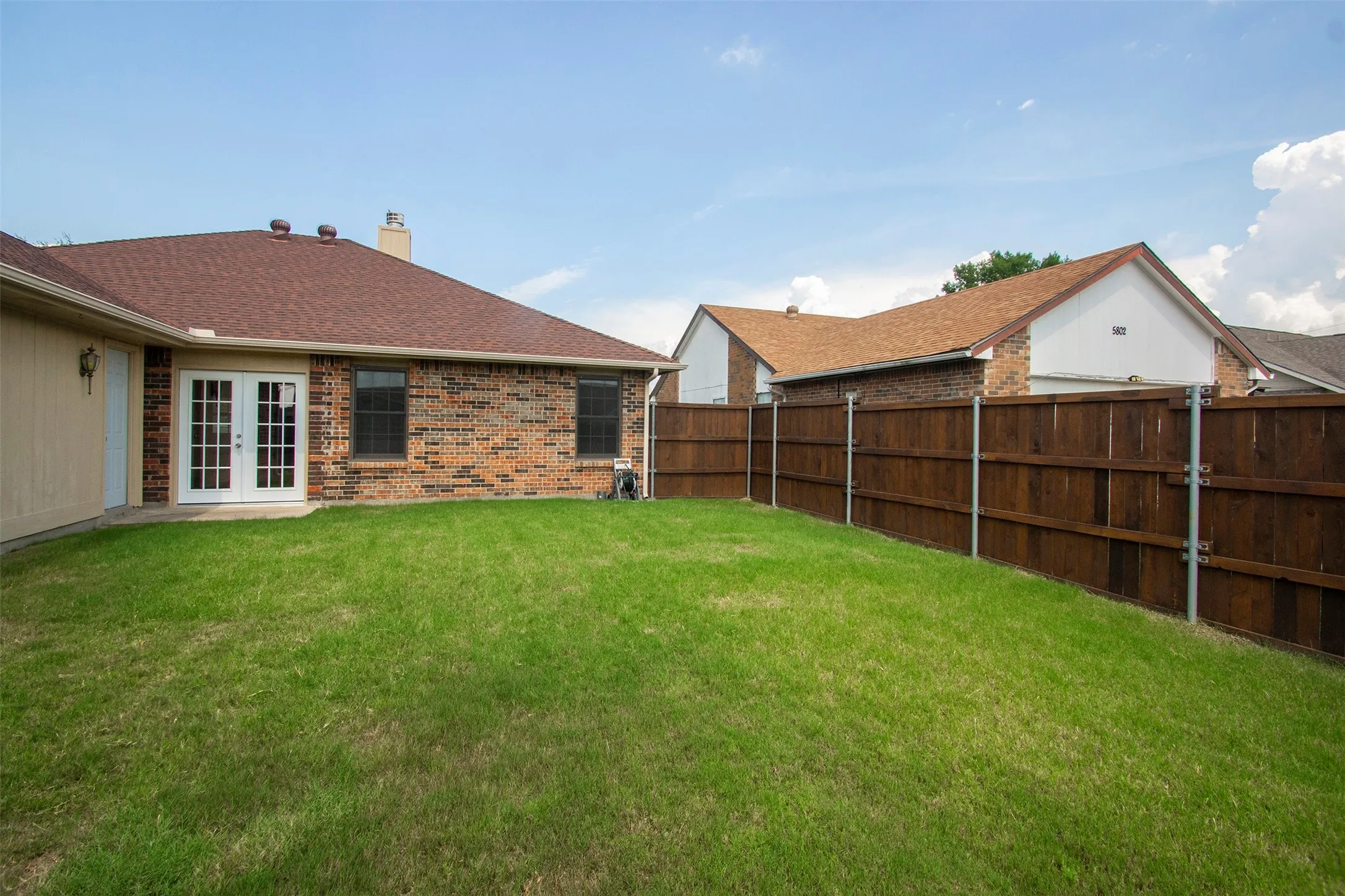 Expansive fenced backyard featuring a manicured lawn, brick exterior, white French doors, and a shingle roof