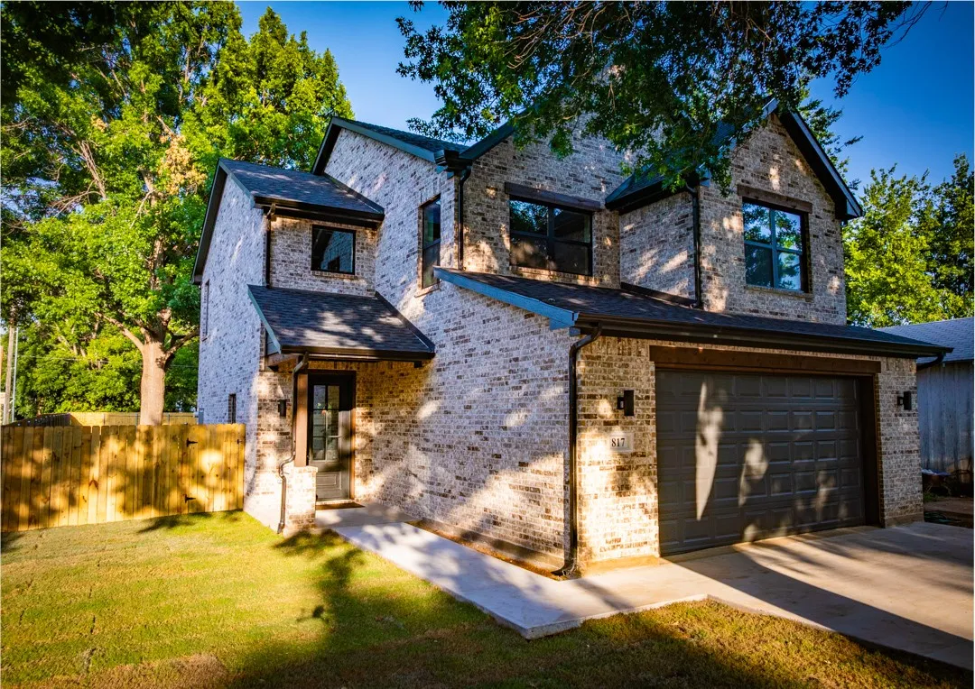 Contemporary two-story brick exterior with a dark gray garage door, black-framed windows, a covered front entry with wood beam accents, and a fenced yard