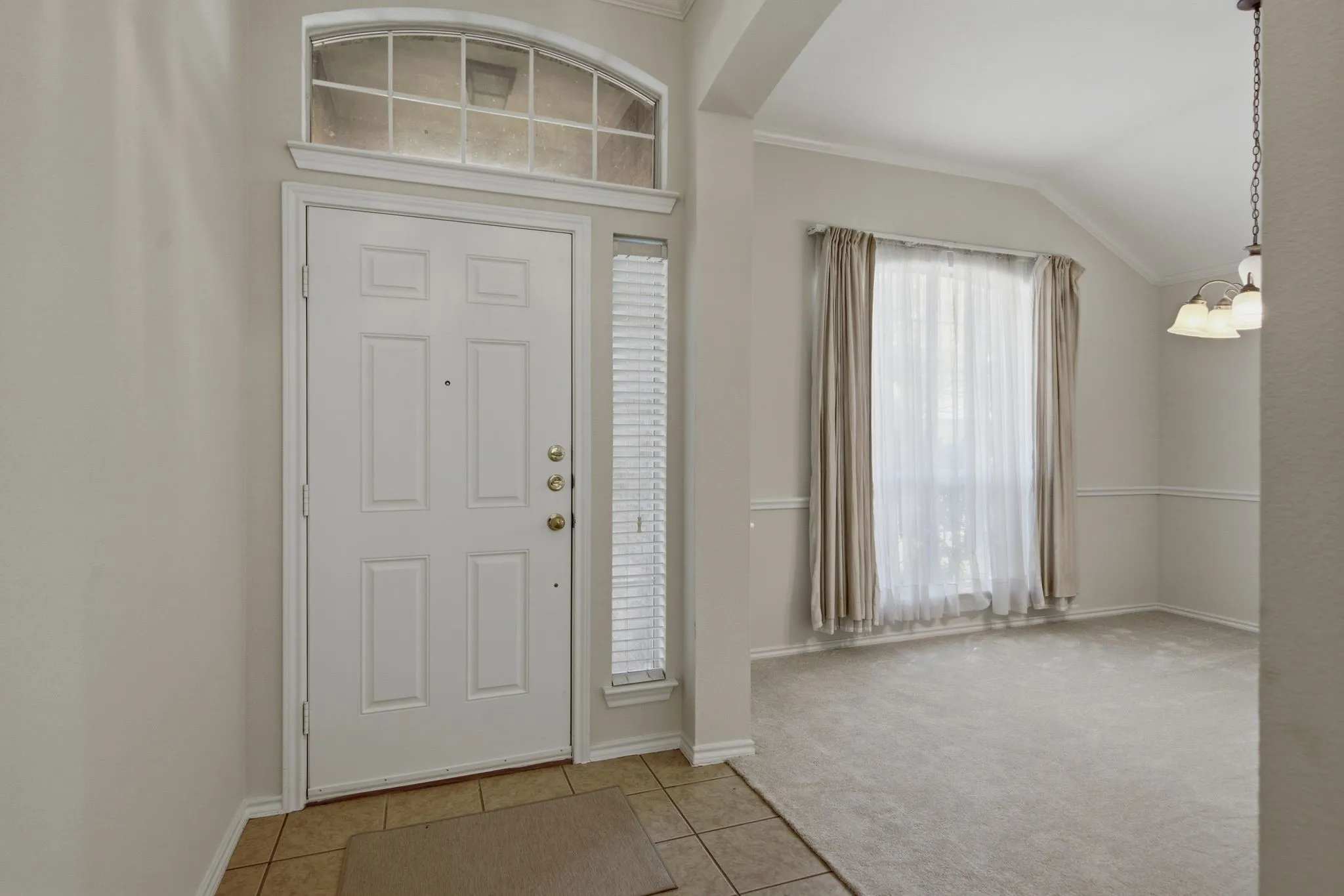 Entryway featuring a paneled door with an arched transom window, a tiled floor, and an adjacent carpeted room with a vaulted ceiling, crown molding, and a chandelier