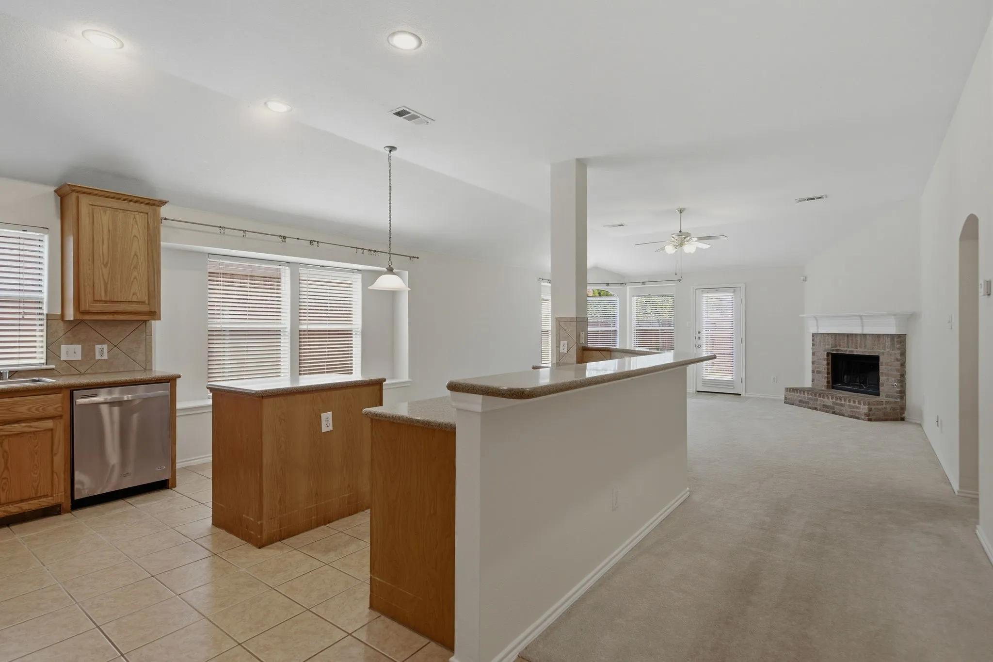 Open-concept kitchen and living area featuring a breakfast bar with a light-toned countertop, wood cabinetry, & stainless steel dishwasher.
