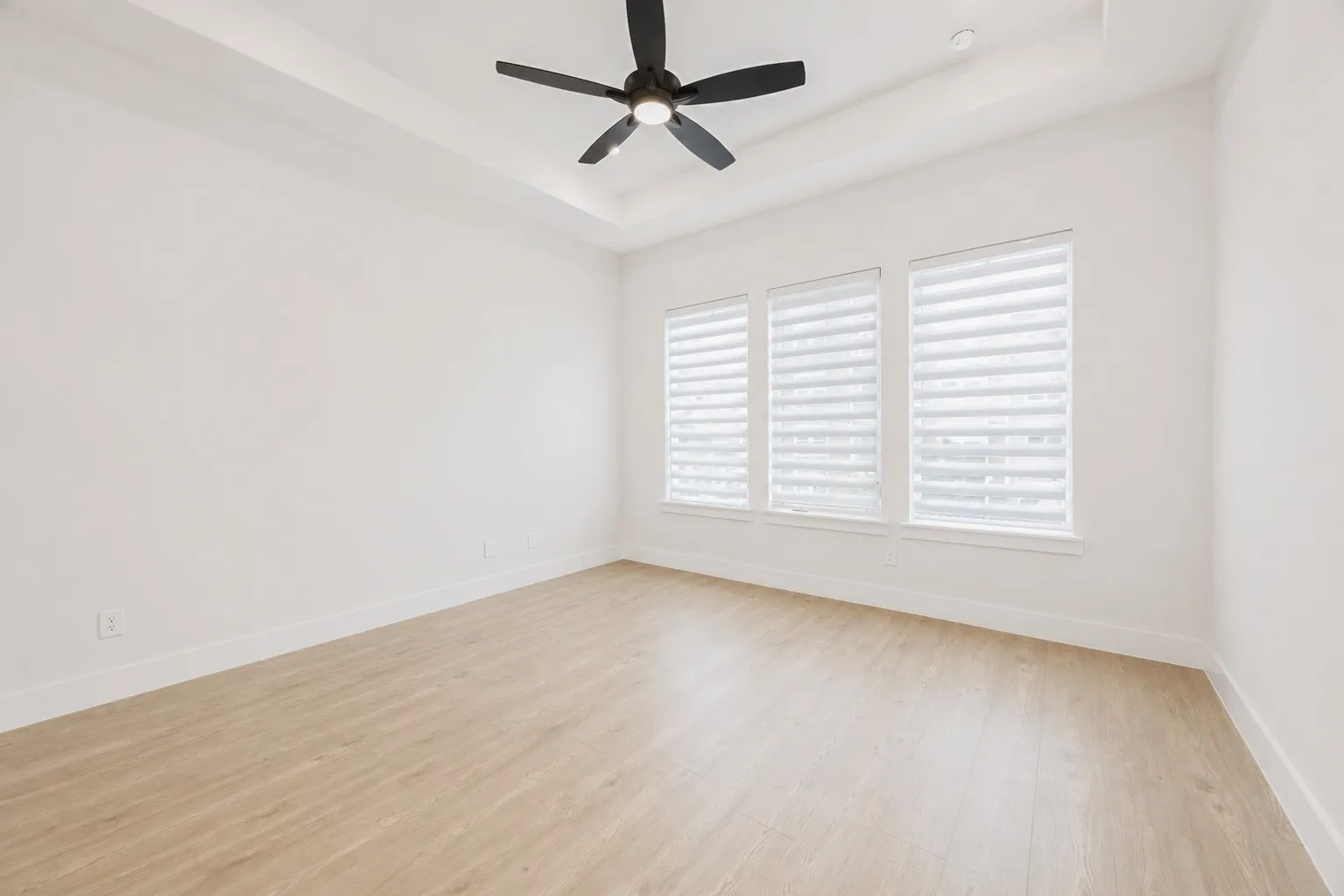 Spacious room featuring wood-finish flooring, a tray ceiling, and a black ceiling fan with integrated lighting