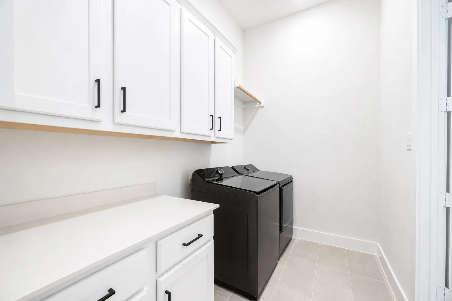 Laundry area featuring white shaker-style cabinetry with black hardware, white countertop space, and light-toned floor tiling