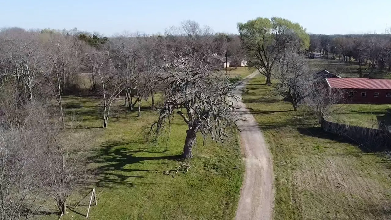 Expansive land parcel featuring a winding dirt road, mature trees, open grassy areas, and a red-roofed barn