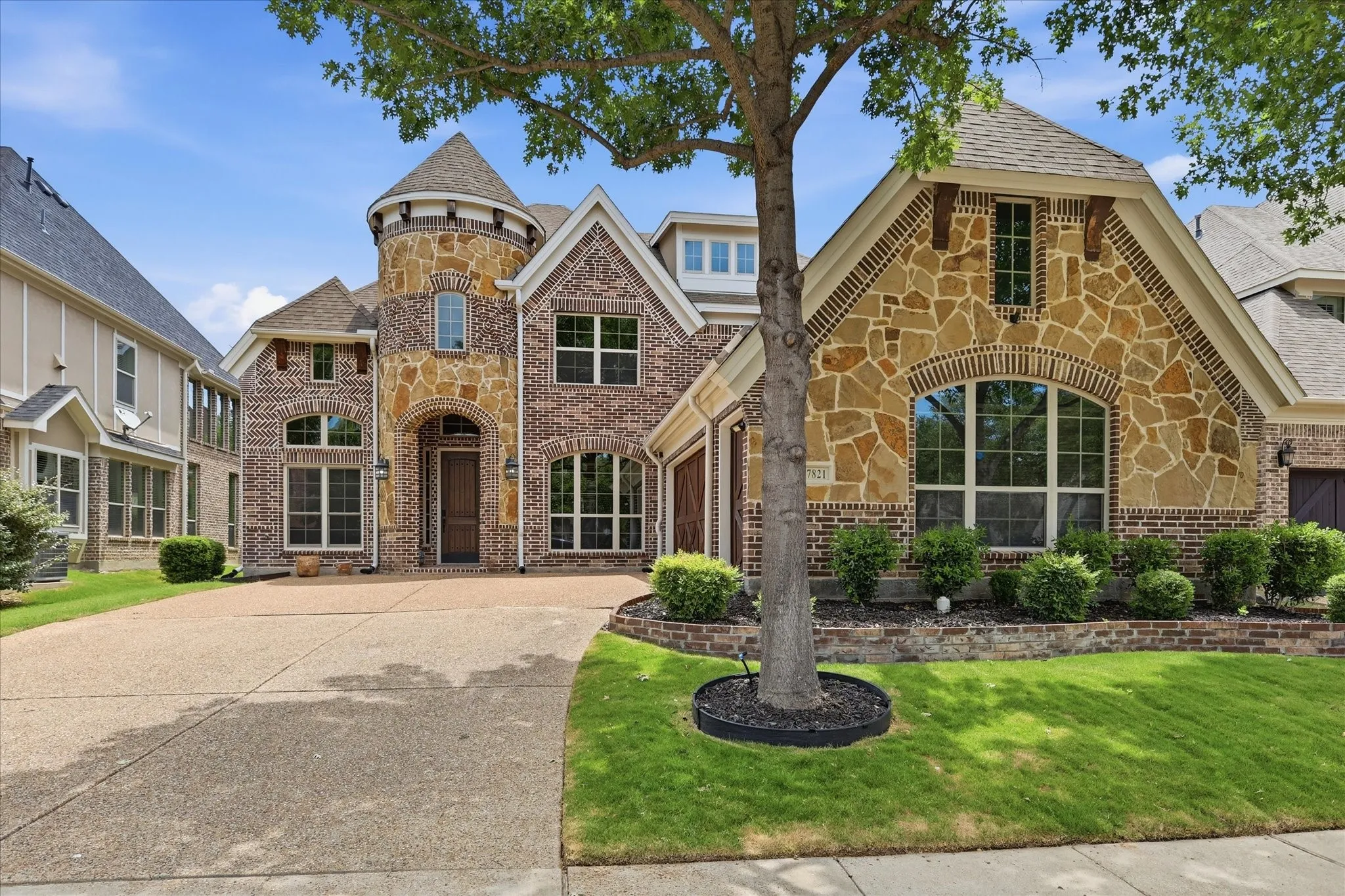 Stunning brick and stone elevation showcasing a dramatic turret with a conical roof, soaring gables, and arched entryways. A charming mix of stacked stone and brick detailing, wood-beam accents, and multi-pane windows create striking curb appeal.