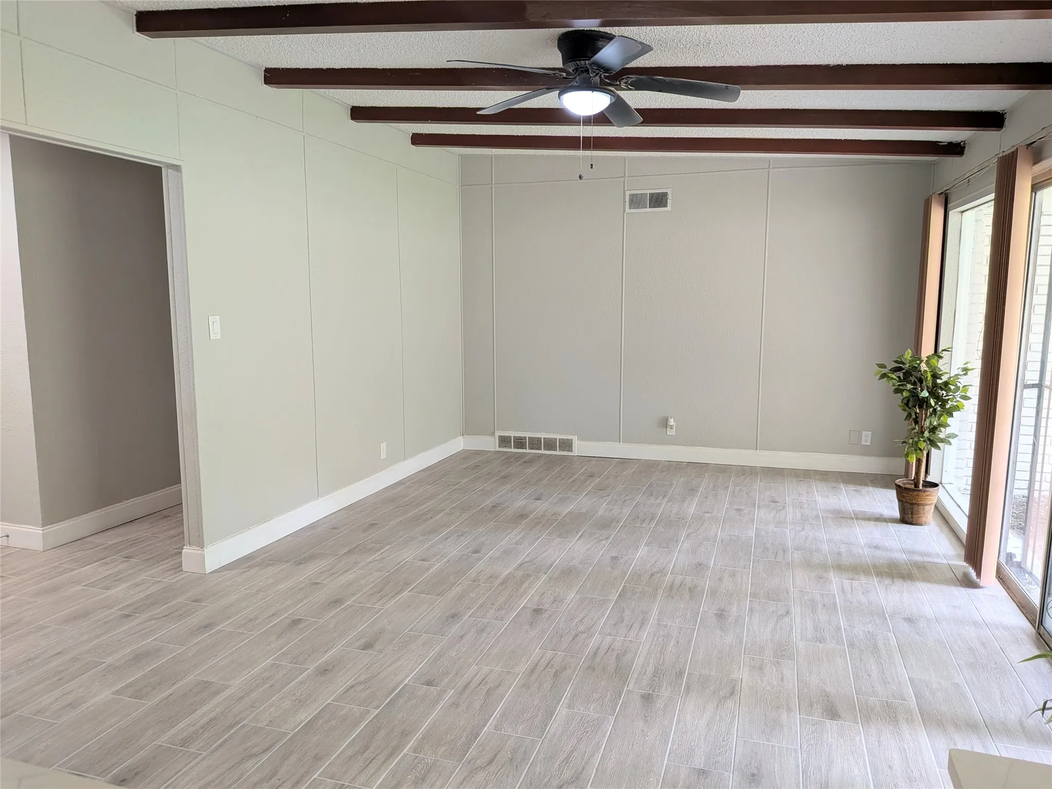 Spacious living area featuring light wood-finish plank flooring, exposed wood ceiling beams, a ceiling fan with integrated lighting, and large windows with wood framing