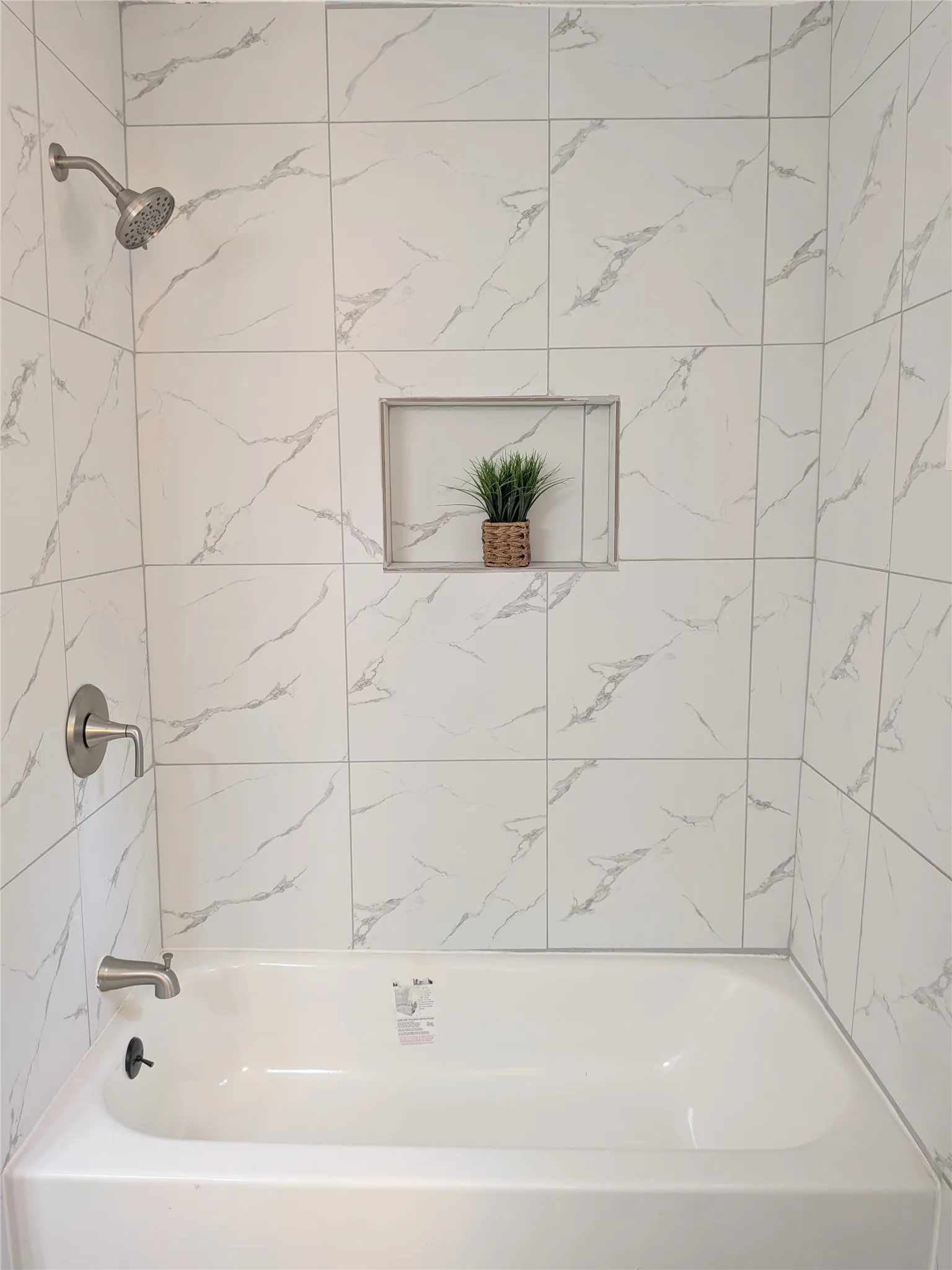 Bathtub and shower combination featuring white tiled walls with subtle gray veining, a brushed nickel shower head, a brushed nickel tub spout, and a recessed shower niche