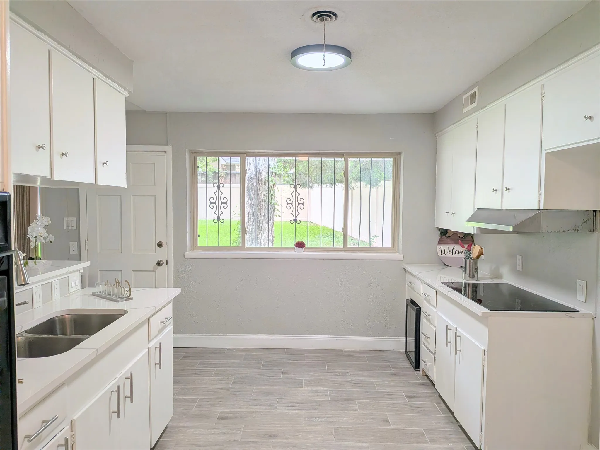 Bright kitchen featuring white cabinetry, light-toned countertops, a stainless steel sink, a built-in cooktop with range hood, and wood-finish tile flooring