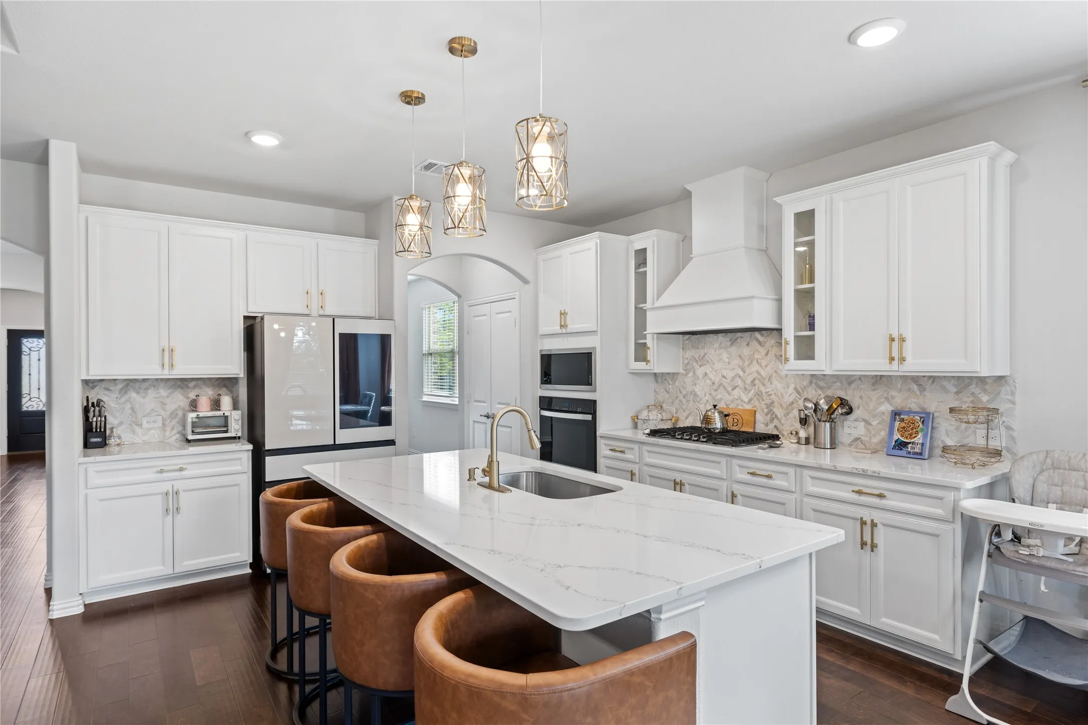 Chef-inspired gourmet kitchen featuring an oversized quartz waterfall island, upgraded vent hood, custom backsplash, gas cooktop, built-in oven and microwave, white shaker cabinetry, designer gold hardware, pendant lighting, and abundant storage.