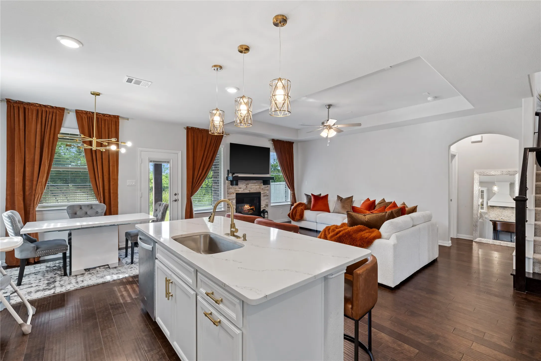 Open-concept living area featuring a kitchen island with sink and gold-finish faucet, a stacked stone fireplace, dark wood-finish flooring, and a tray ceiling. Expansive open-concept kitchen overlooking the spacious family room with fireplace, abundant natural light, oversized island seating, breakfast area, and seamless indoor-outdoor flow to the extended covered patio.
