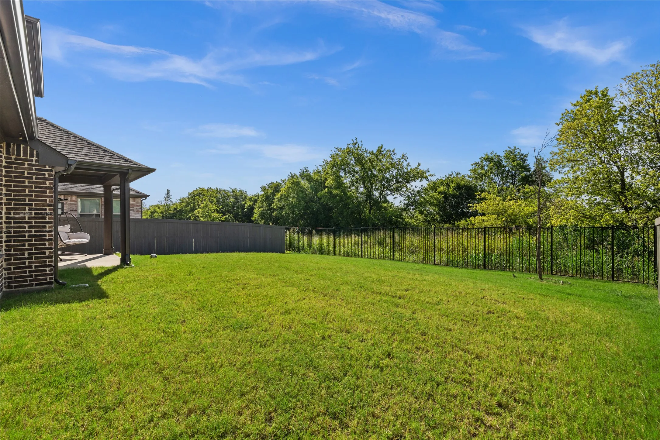 Expansive green lawn with a covered patio, brick exterior, and a combination privacy and wrought iron fencing. Spacious fenced backyard featuring a covered patio and premium greenbelt lot near rear neighbors. Ideal for outdoor entertaining, family, gatherings, gardening, or relaxing while enjoying scenic views and added privacy.