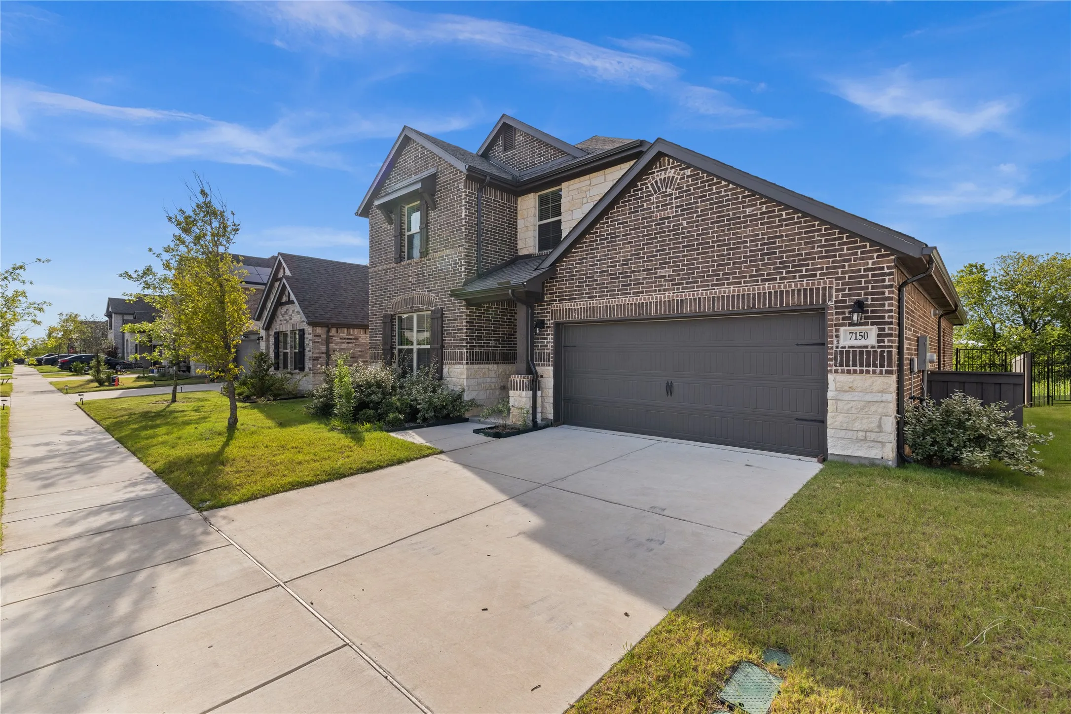 Two-story residence featuring a brick and stone facade, dark-toned siding, a two-car garage, and a concrete driveway. Corner perspective showcasing the home’s elegant architecture, extended driveway, mature landscaping, upgraded exterior lighting, and inviting covered entry. Beautiful streetscape with sidewalks and modern neighboring homes.
