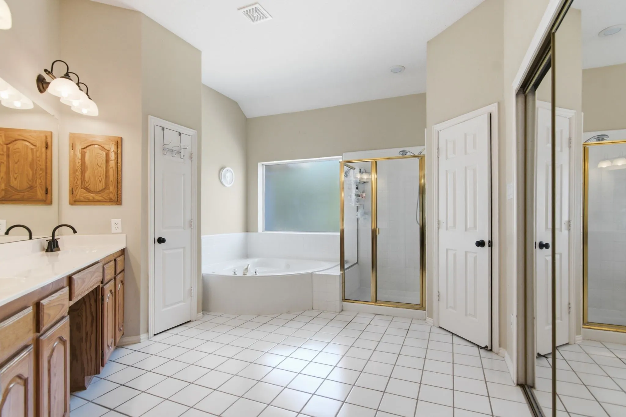 Expansive bathroom featuring a corner soaking tub, separate shower with brass-finish enclosure, dual vanity with wood cabinetry and white countertop, and large walk-in closet.