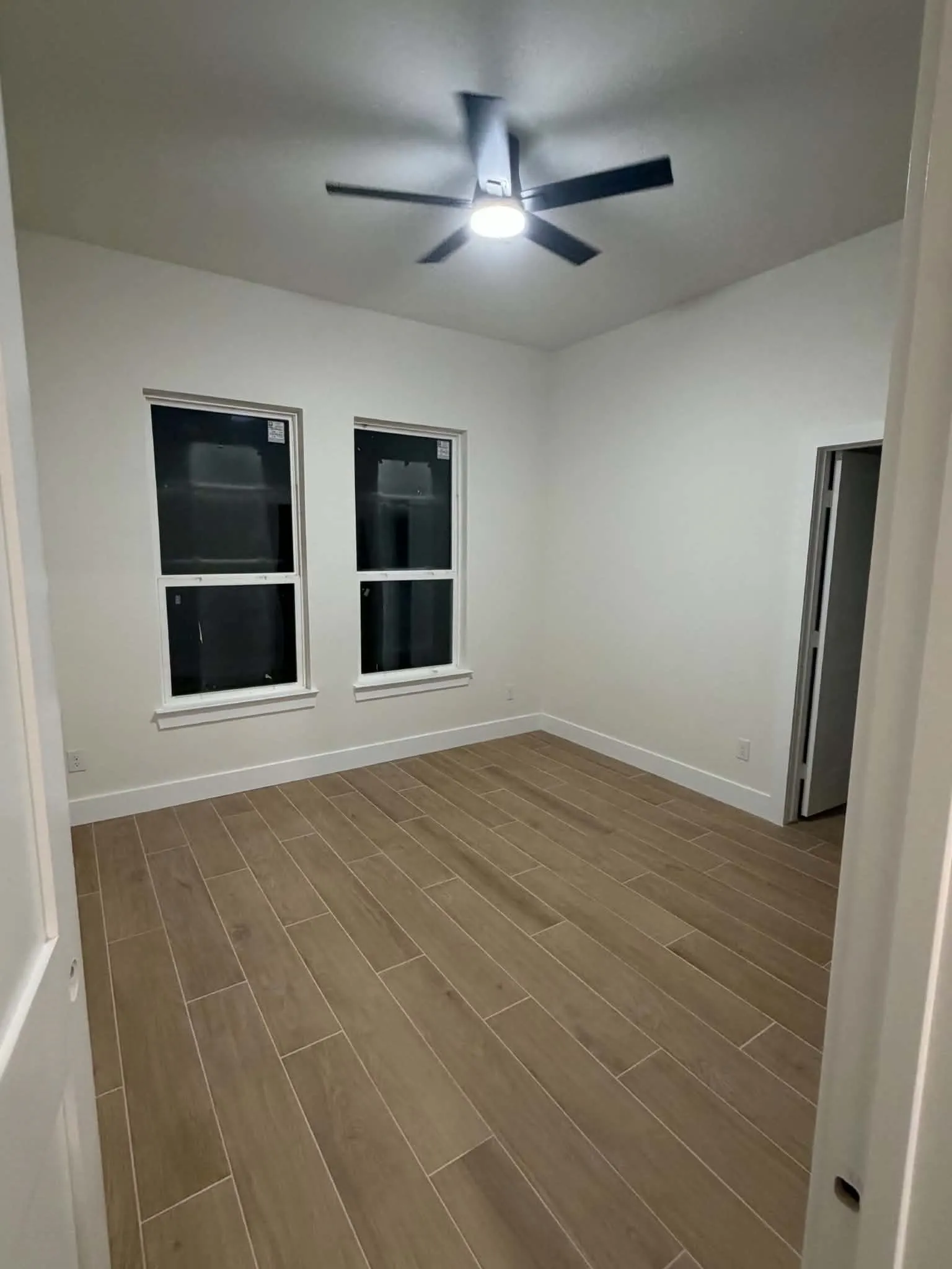 Wood-finish plank flooring throughout, featuring a ceiling fan with integrated lighting, and two windows with white trim