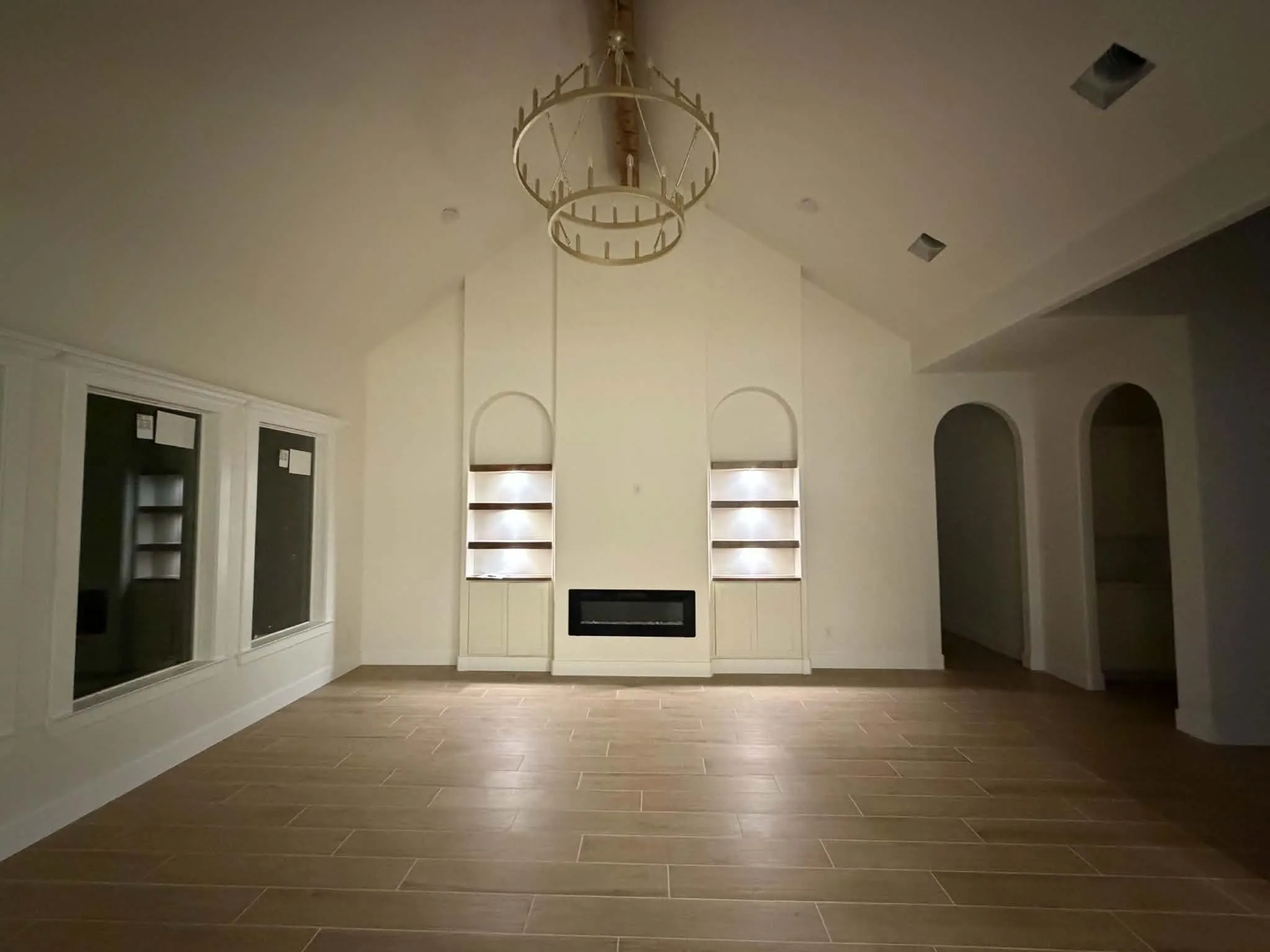 Vaulted ceiling with an ornate chandelier, built-in fireplace, two arched built-in shelving units with integrated lighting, and wood-finish flooring