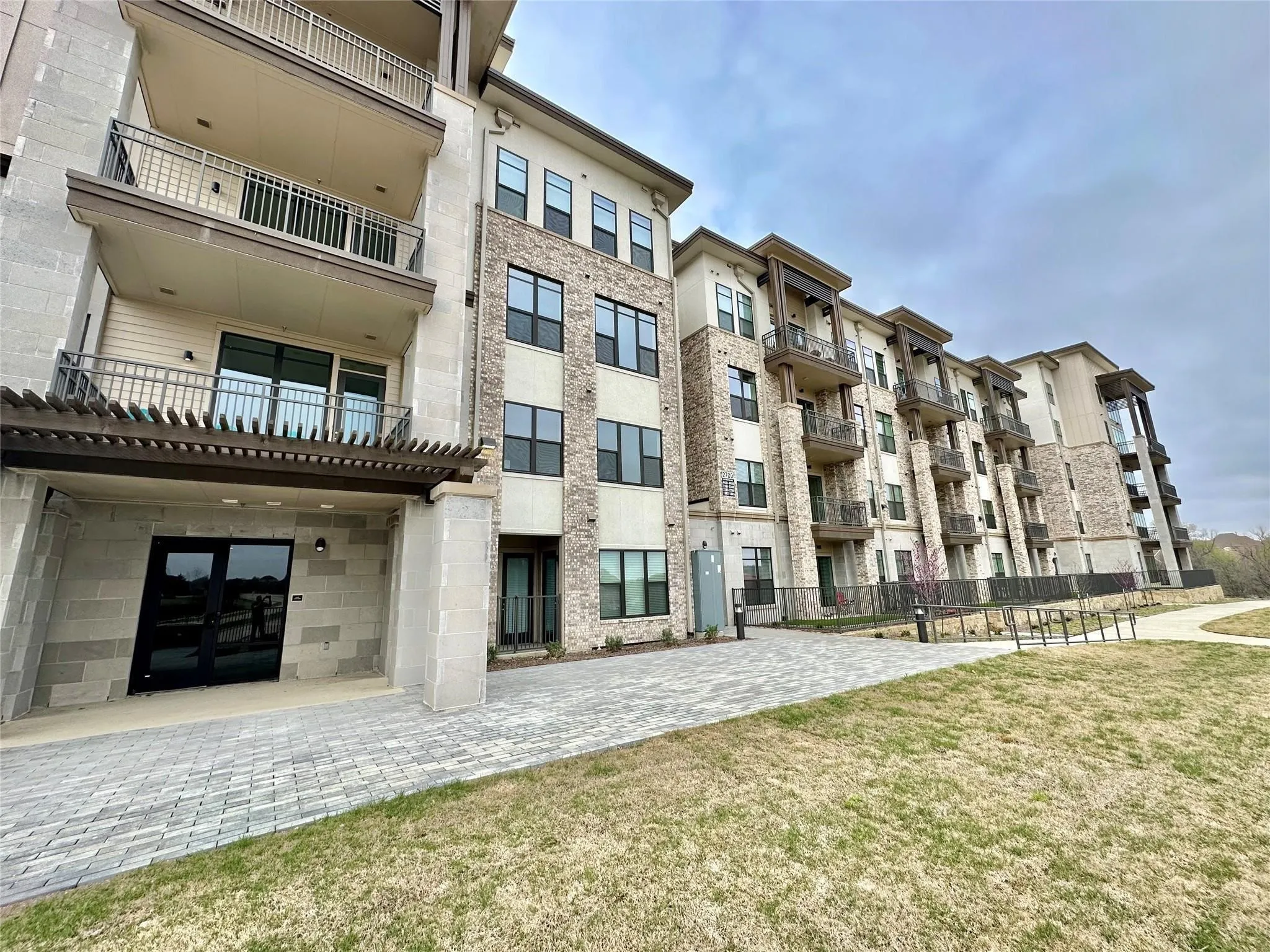 Multi-story building featuring stone and brick façade accents, private balconies with metal railings, dark-framed windows, a ground-level entrance with double glass doors, and a decorative pergola