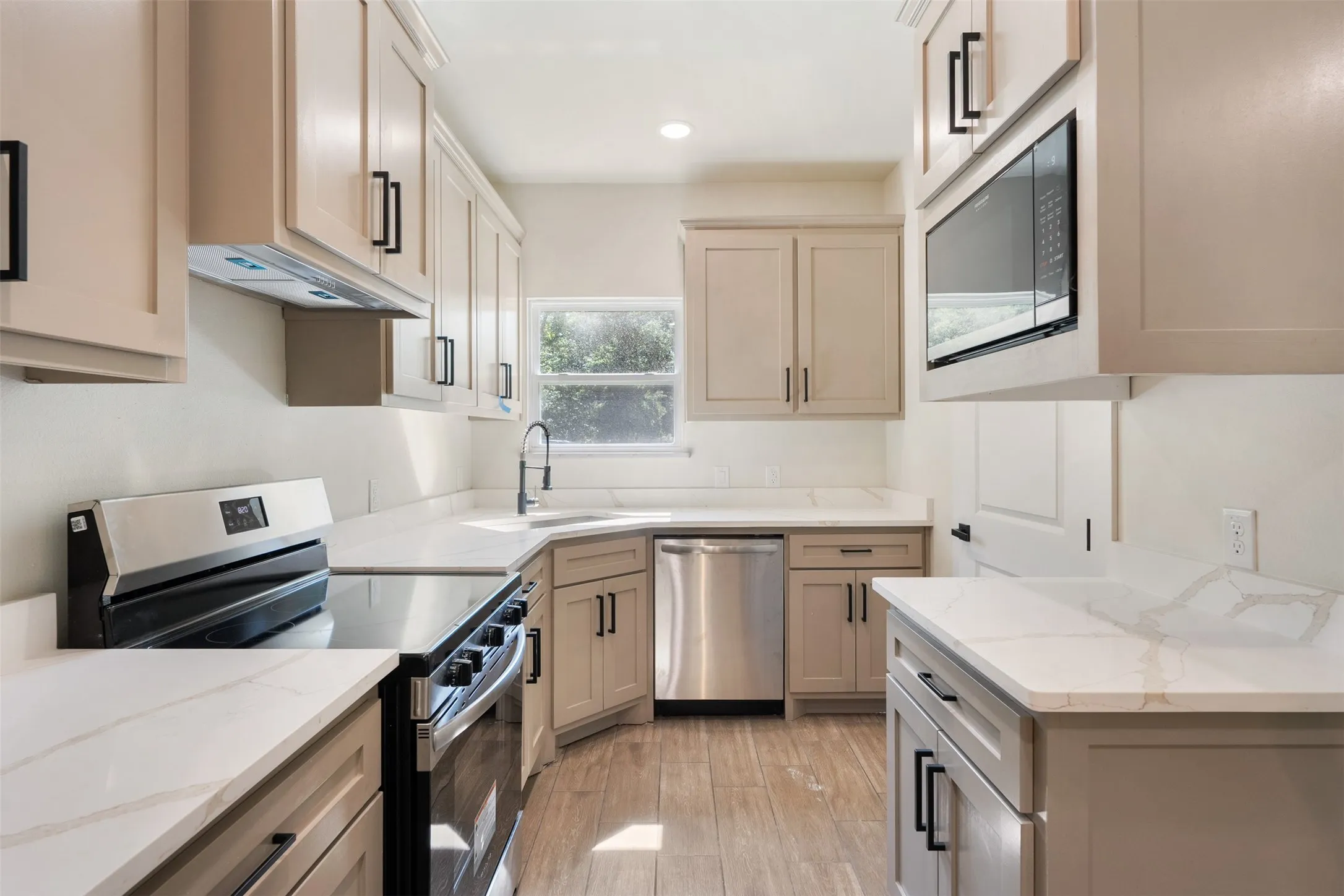 Contemporary kitchen featuring white solid surface countertops, stainless steel appliances, wood-finish flooring, light-toned cabinetry, and black hardware