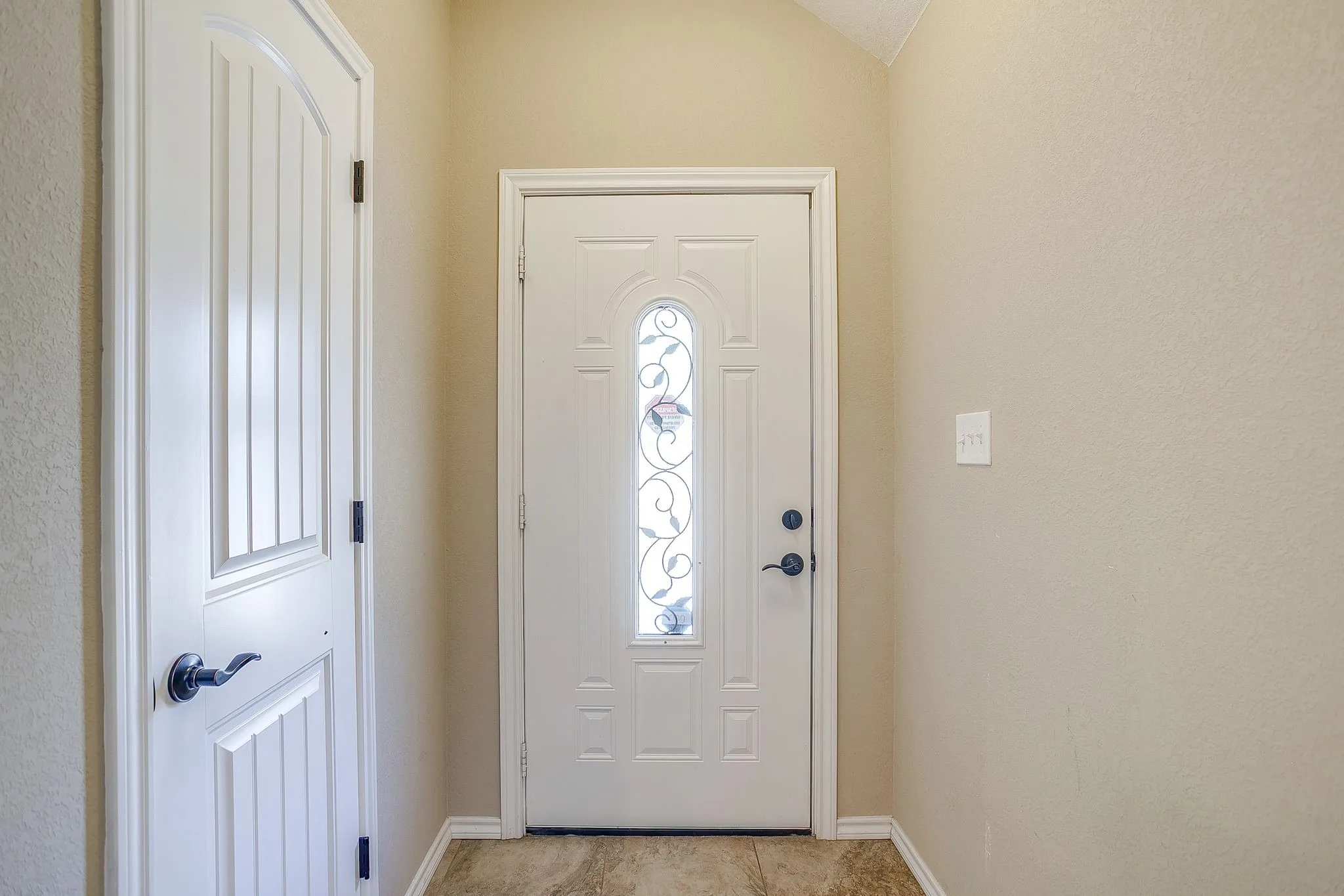 Entryway featuring a white paneled door with decorative glass inlay, textured light beige walls, and brown tile flooring