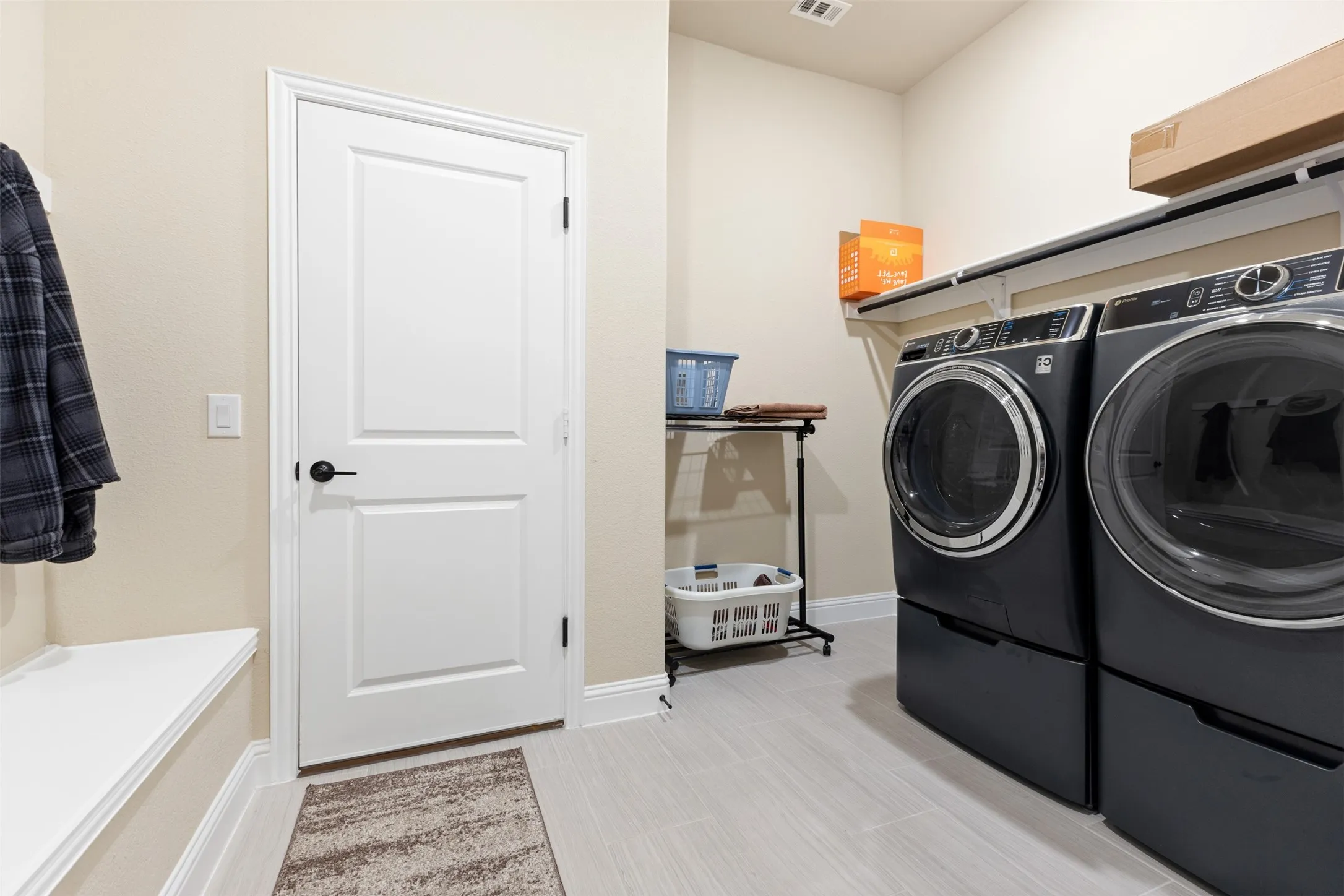 Dedicated laundry area featuring modern front-load appliances on pedestals, overhead shelving, and light-toned tile flooring