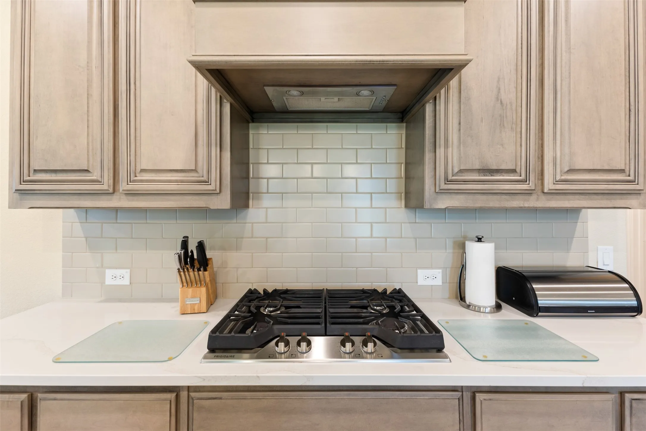 Kitchen featuring a stainless steel gas cooktop with four burners, a built-in range hood, light gray subway tile backsplash, light wood-finish cabinetry, and solid surface countertops