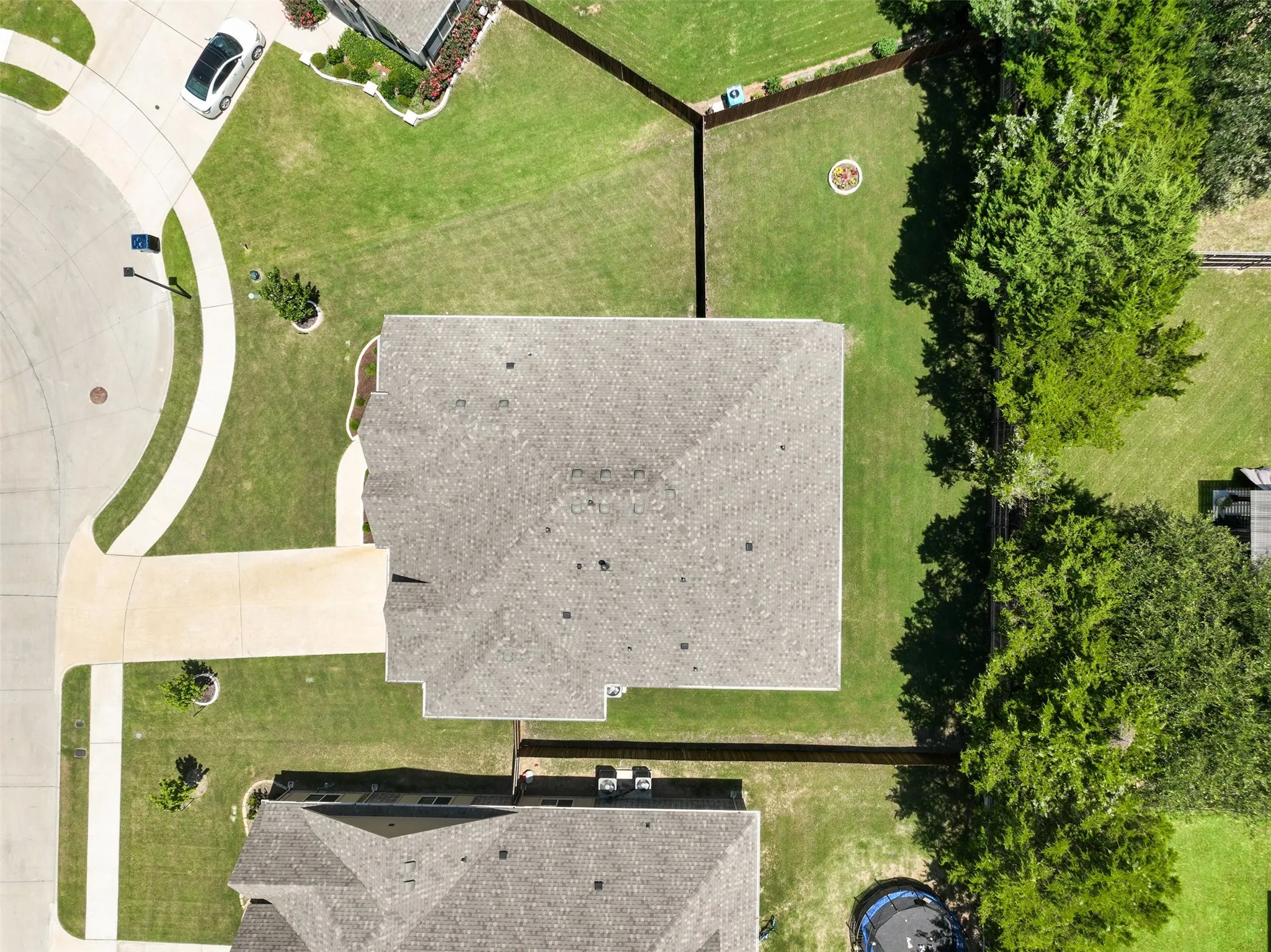 Aerial perspective showcasing a property with a grey shingle roof, extensive green lawn, concrete driveway, and mature trees bordering the rear perimeter