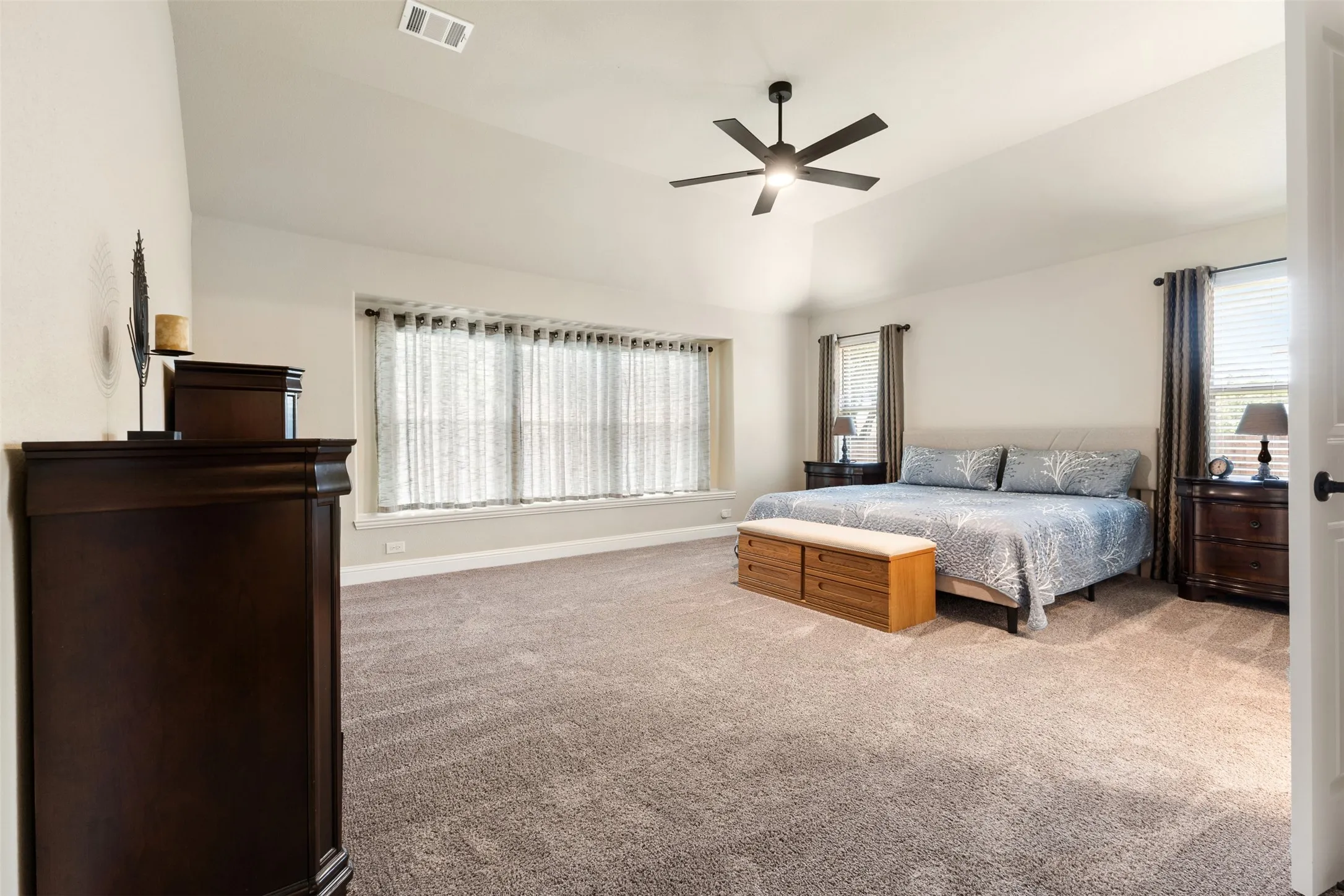 Spacious bedroom featuring vaulted ceilings, neutral-toned carpeting, a contemporary ceiling fan, and multiple windows with blinds
