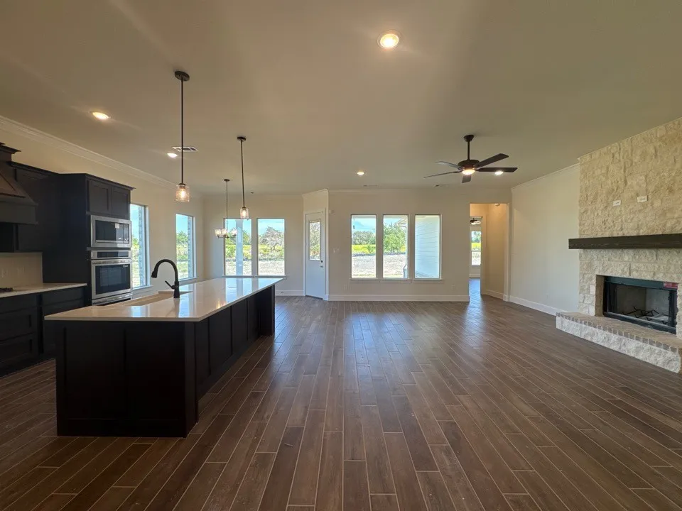 Open-concept living space with wood-finish flooring, a stone fireplace with a wood mantle, and a kitchen island with pendant lighting