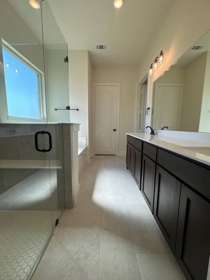 Dual vanity with dark wood-finish cabinetry, frameless glass shower enclosure, built-in shower bench, overhead recessed lighting, and large format tile flooring