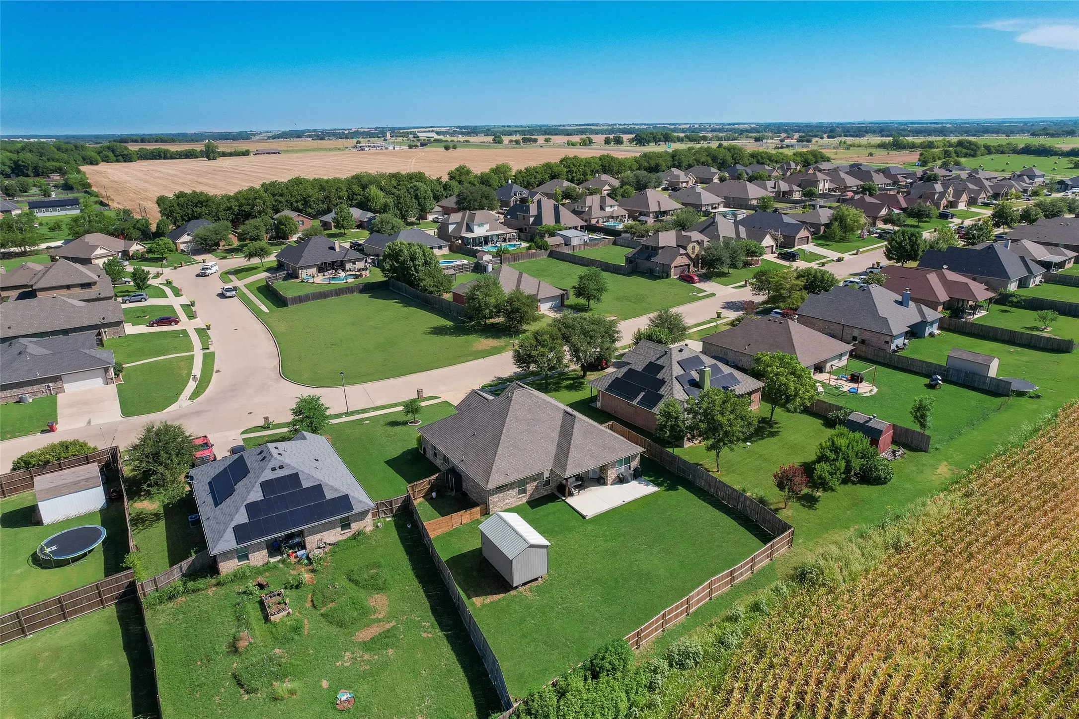 Aerial neighborhood panorama featuring a residential street, manicured lawns, and surrounding agricultural fields