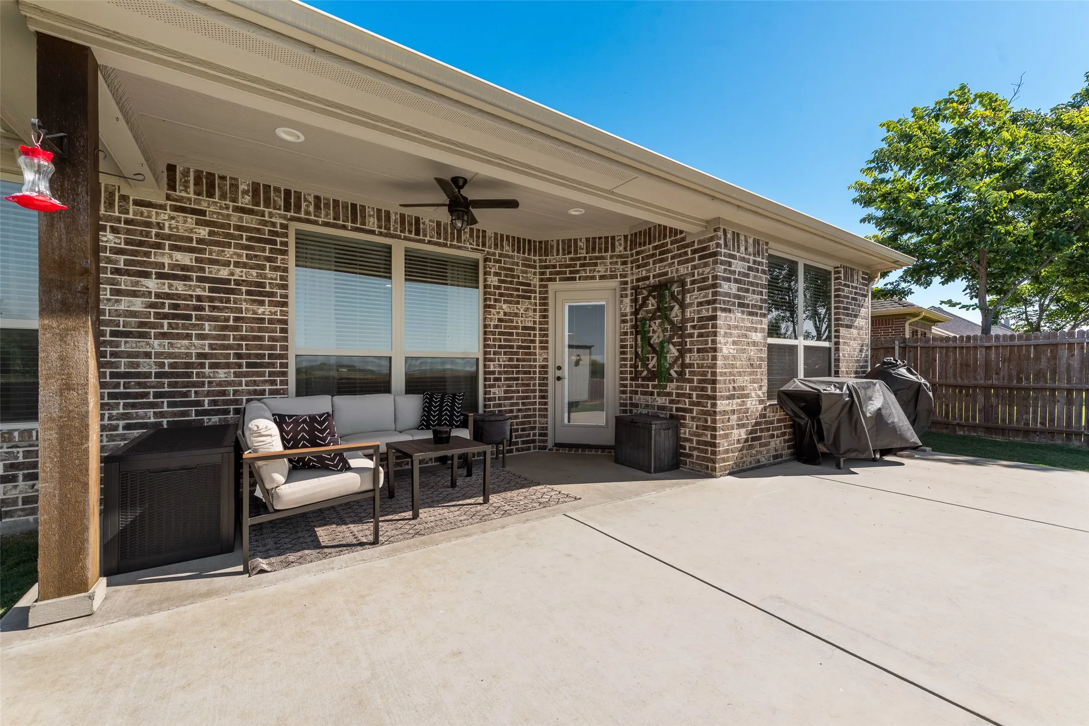 Covered outdoor patio featuring a ceiling fan, brick exterior, multiple windows with blinds, and a glass-paneled door