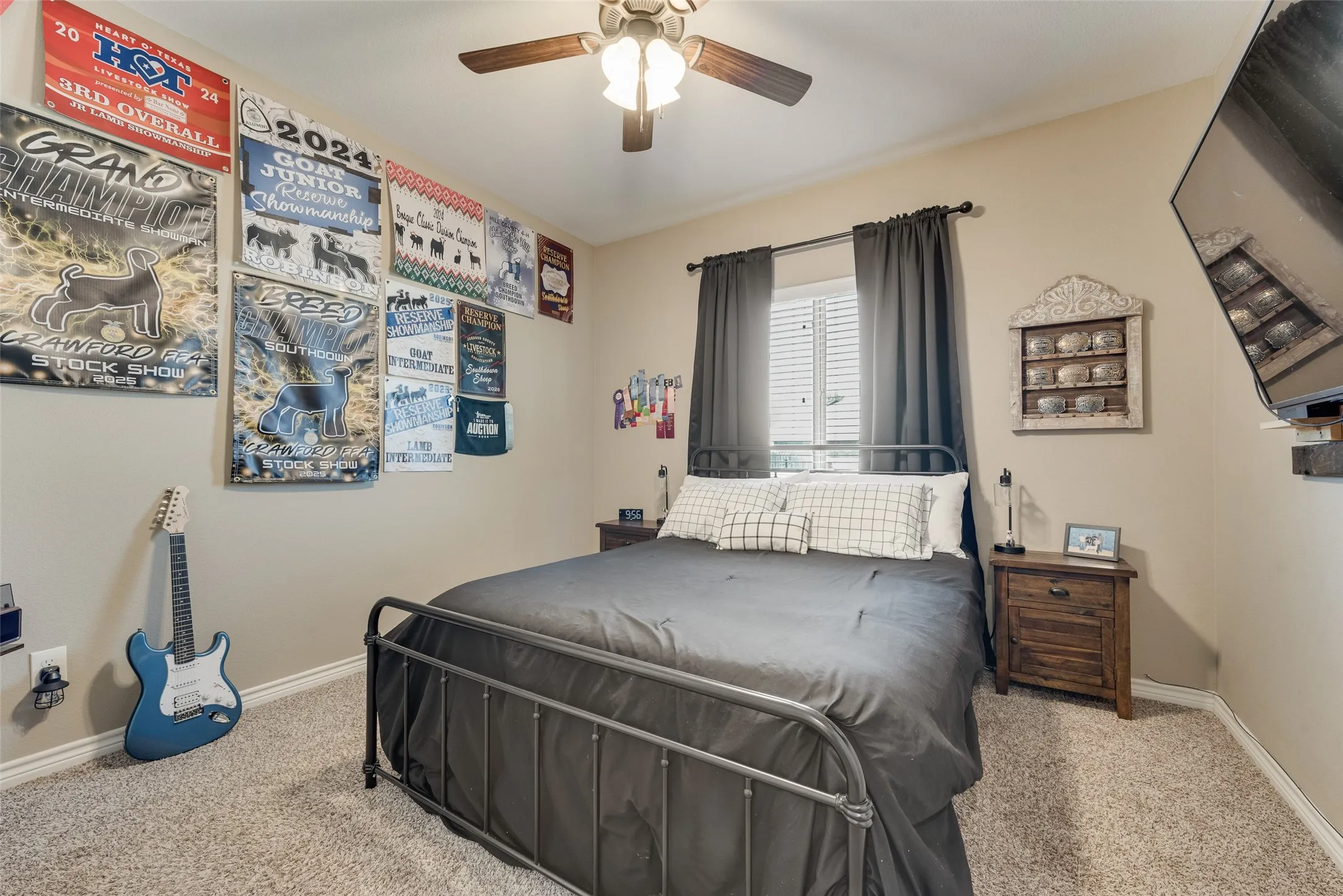 Carpeted room featuring a ceiling fan with light fixture, a window with blinds and dark curtains, and a wood-finish nightstand
