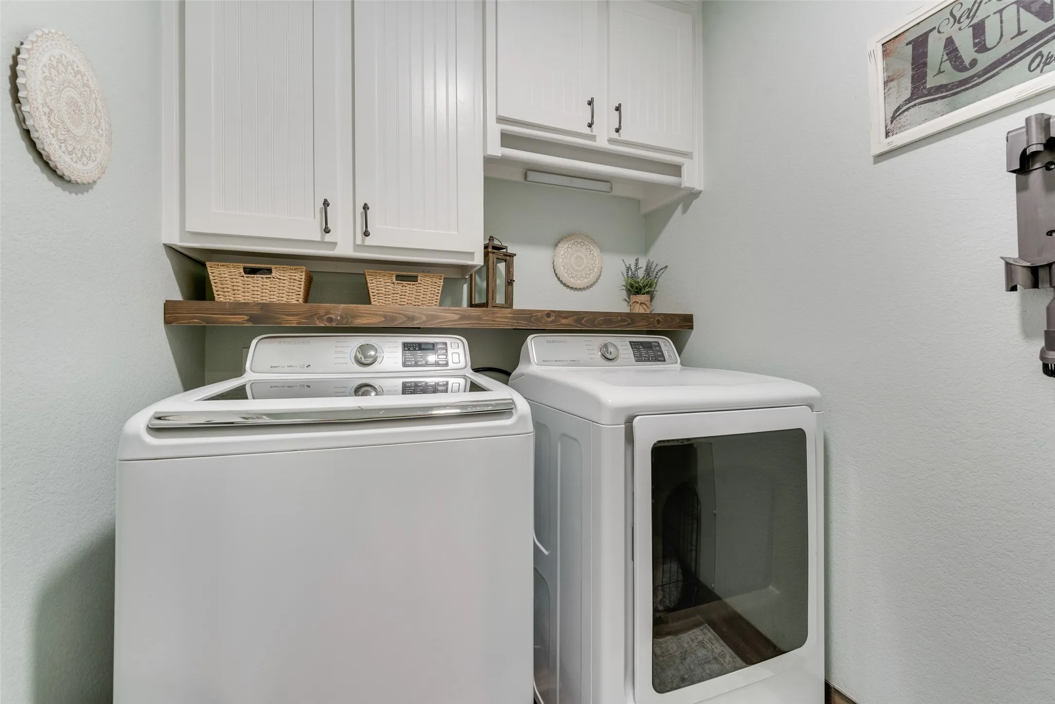 Dedicated laundry space featuring white shaker-style cabinetry, a dark wood-finish floating shelf, and light-toned wall paint