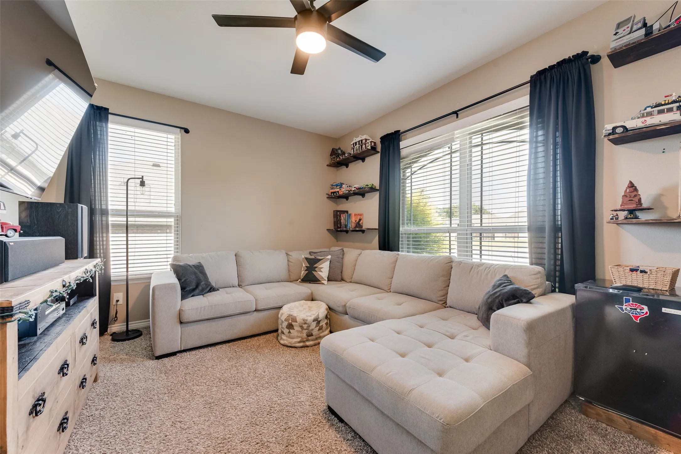 Carpeted room featuring a ceiling fan, two windows with blinds and dark curtains, and wall-mounted shelving